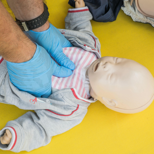 A person in blue gloves practicing infant CPR on a baby mannequin lying on a yellow surface.