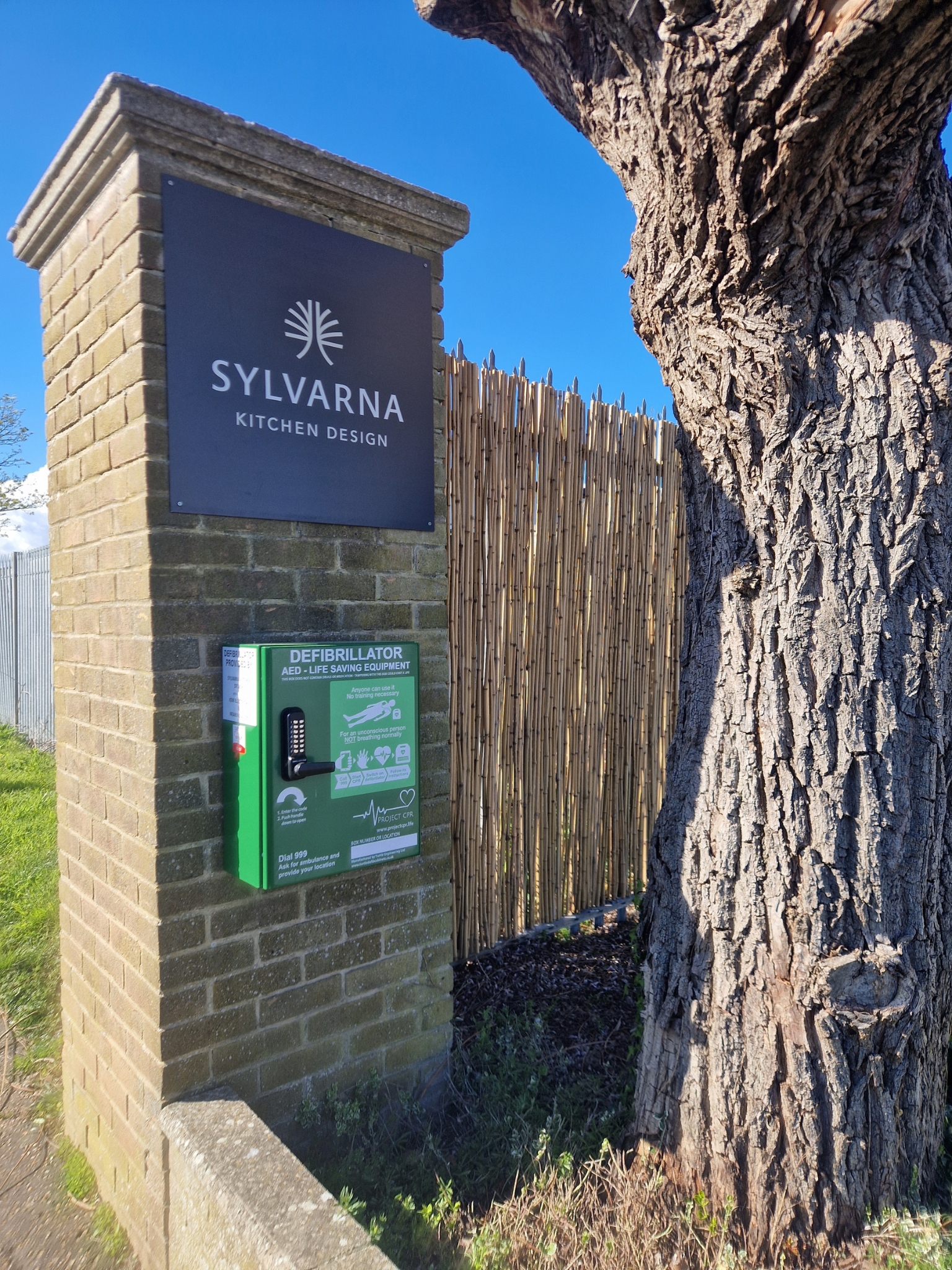 A brick pillar with a sign reading 'Sylvarna Kitchen Design' and a green AED/Defibrillator box mounted underneath, set beside a tree and a bamboo fence under a clear blue sky.