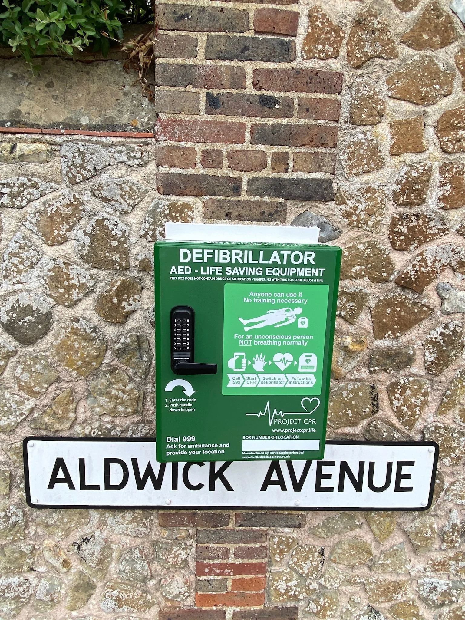 Close-up of a green public access AED cabinet mounted on a stone wall, with a white street name sign below reading 'Aldwick Avenue' and a small part of another sign beneath it.