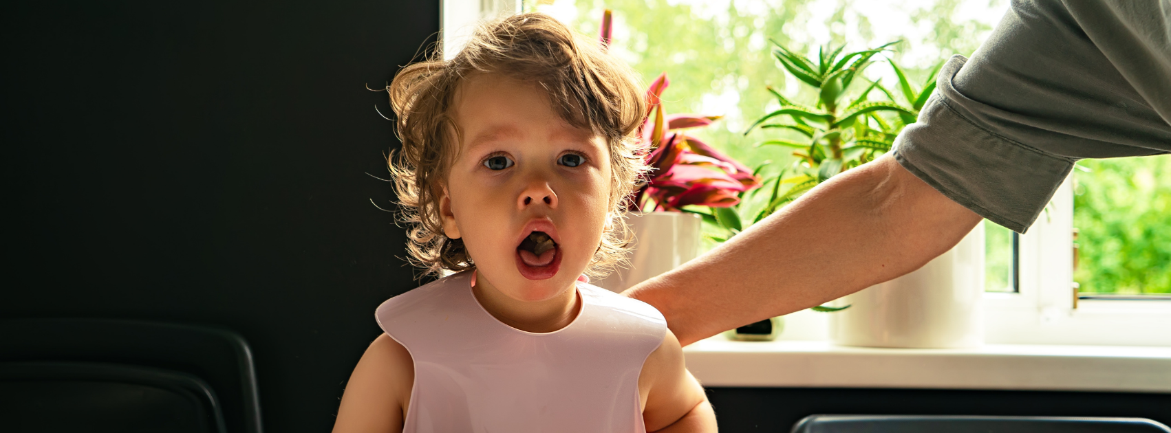 A young child with curly hair standing indoors near a window with green plants, mouth open, possibly crying or talking, with an arm of an adult reaching towards the child.