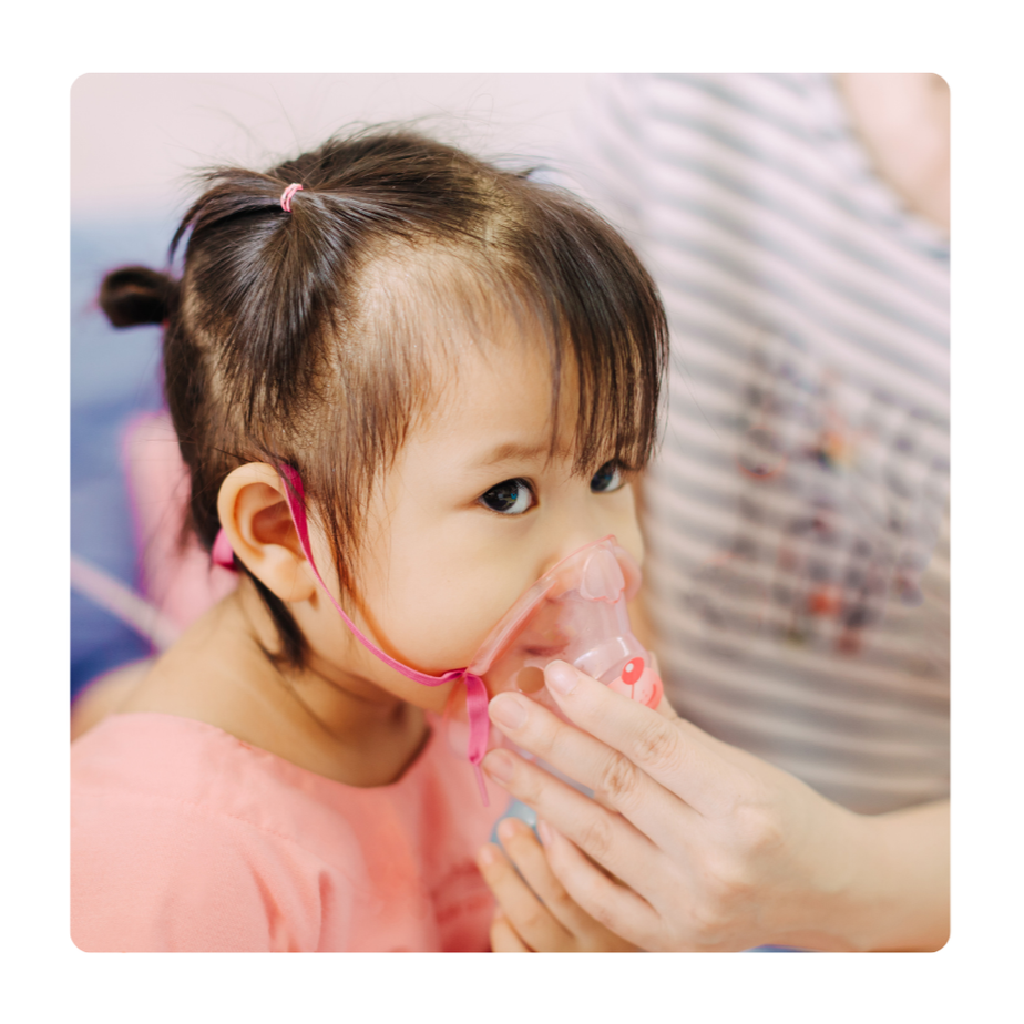 A young girl with dark hair in pigtails wearing a pink dress using a nebulizer with a healthcare provider assisting.