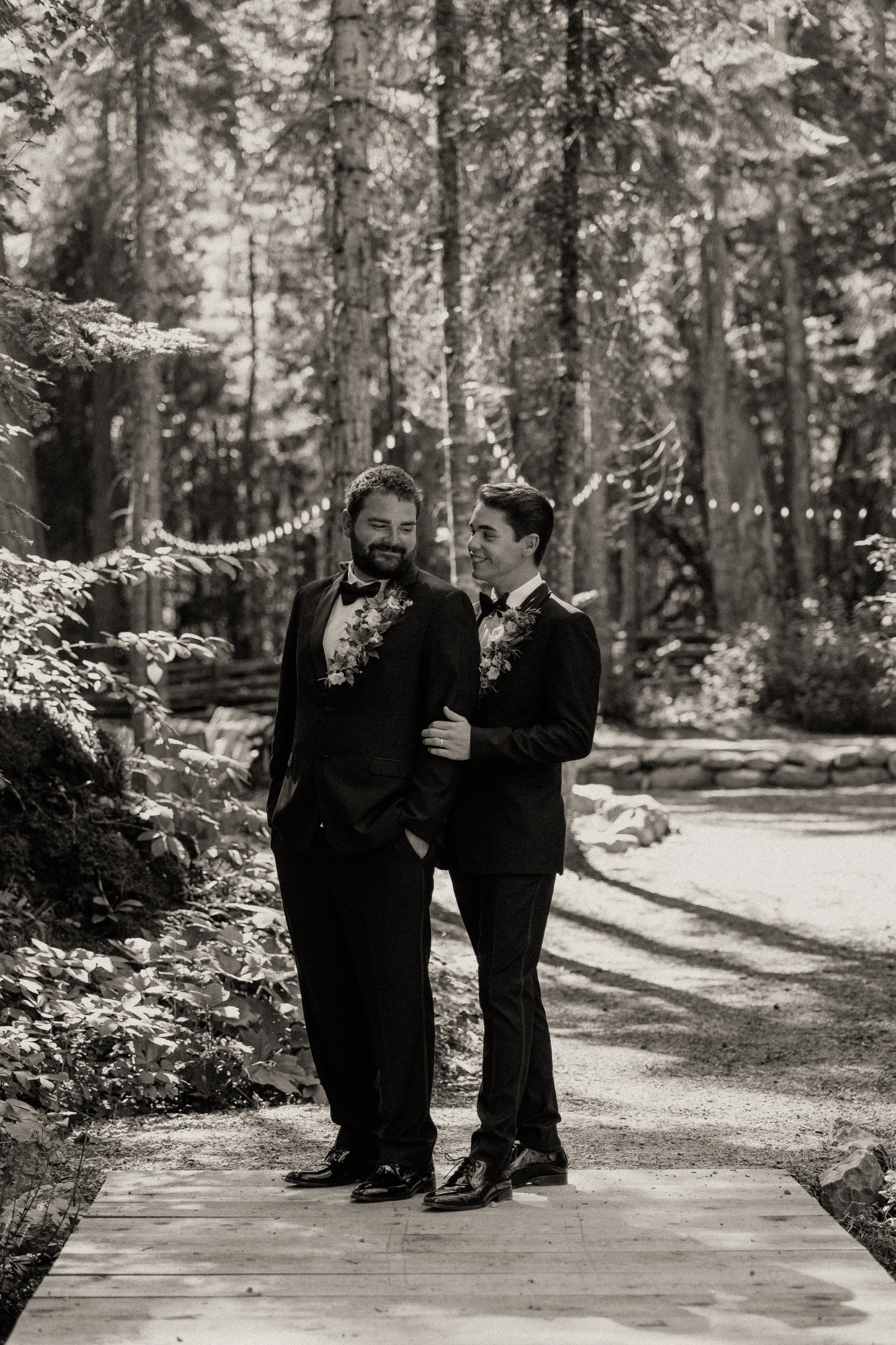 Two men in tuxedos with floral leis, standing together on a wooden bridge in a forest, smiling at each other.