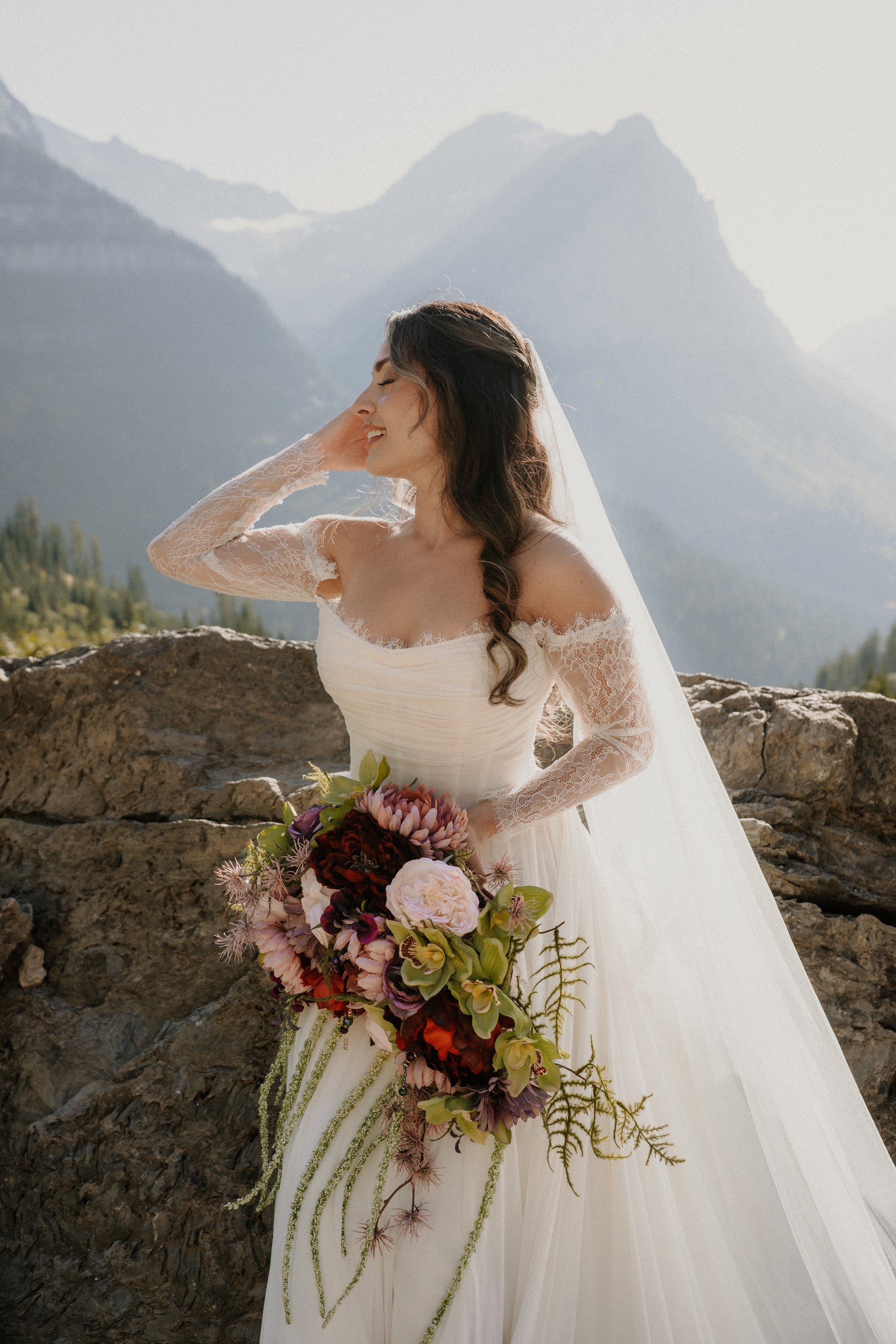 A bride in a white wedding dress with lace sleeves, holding a large bouquet of pink, purple, and green flowers, standing outdoors near rocks with mountains in the background.