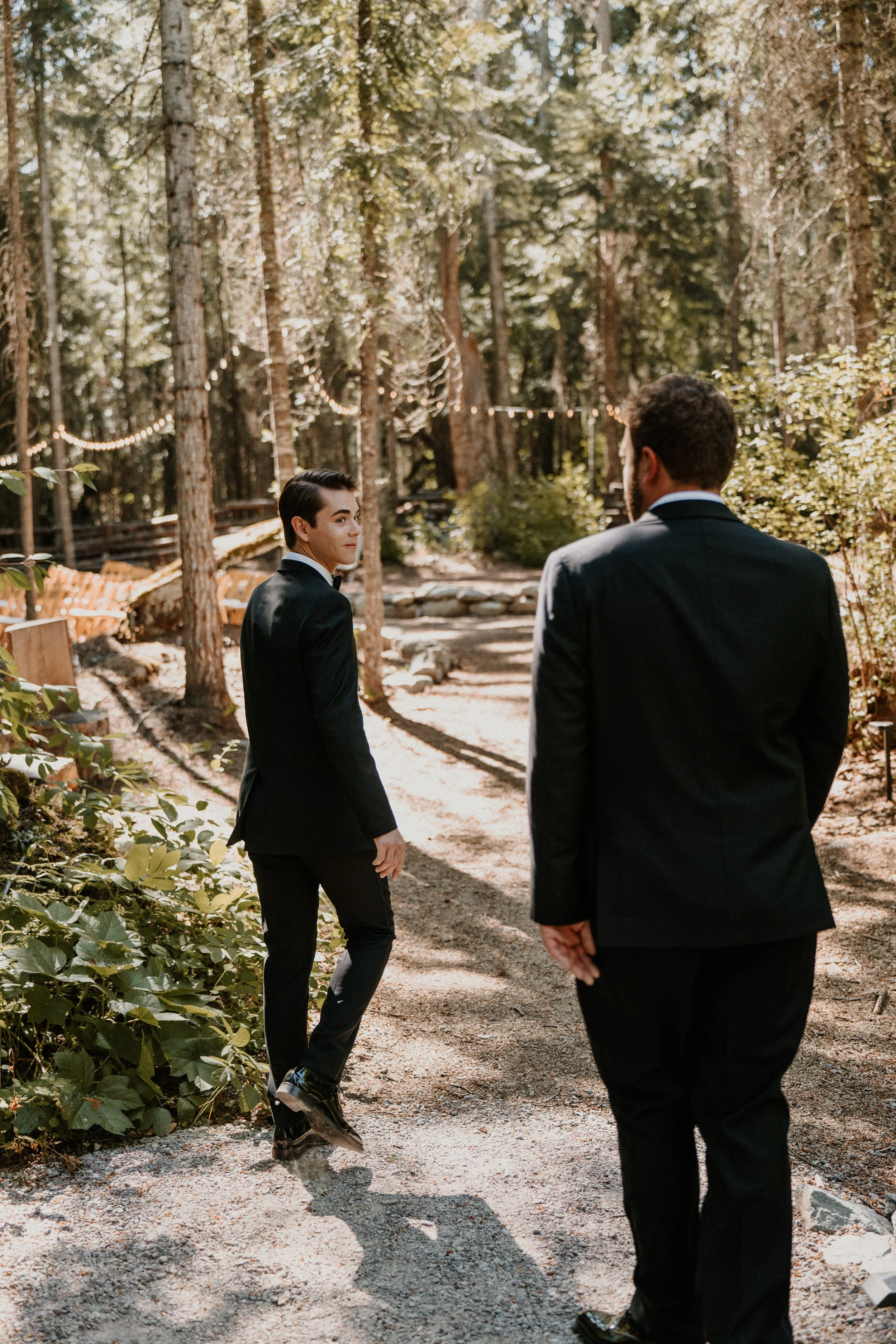 Two men in black suits walking along a forest trail with string lights overhead and natural greenery surrounding them.