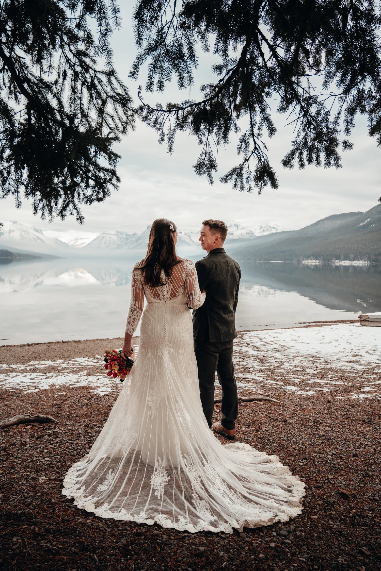 Couple standing together by the lake surrounded by snowy trees and mountains in Glacier National Park