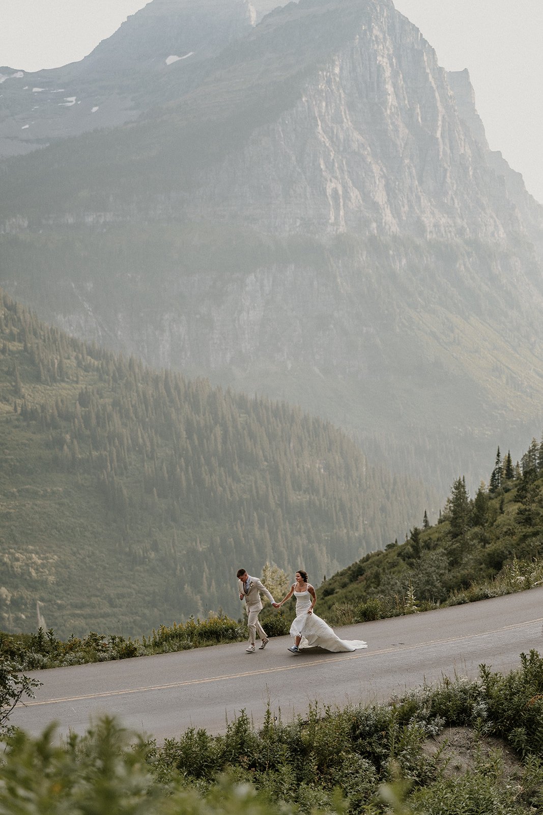 A bride and groom holding hands and walking on a winding mountain road with tall, forested mountains in the background.