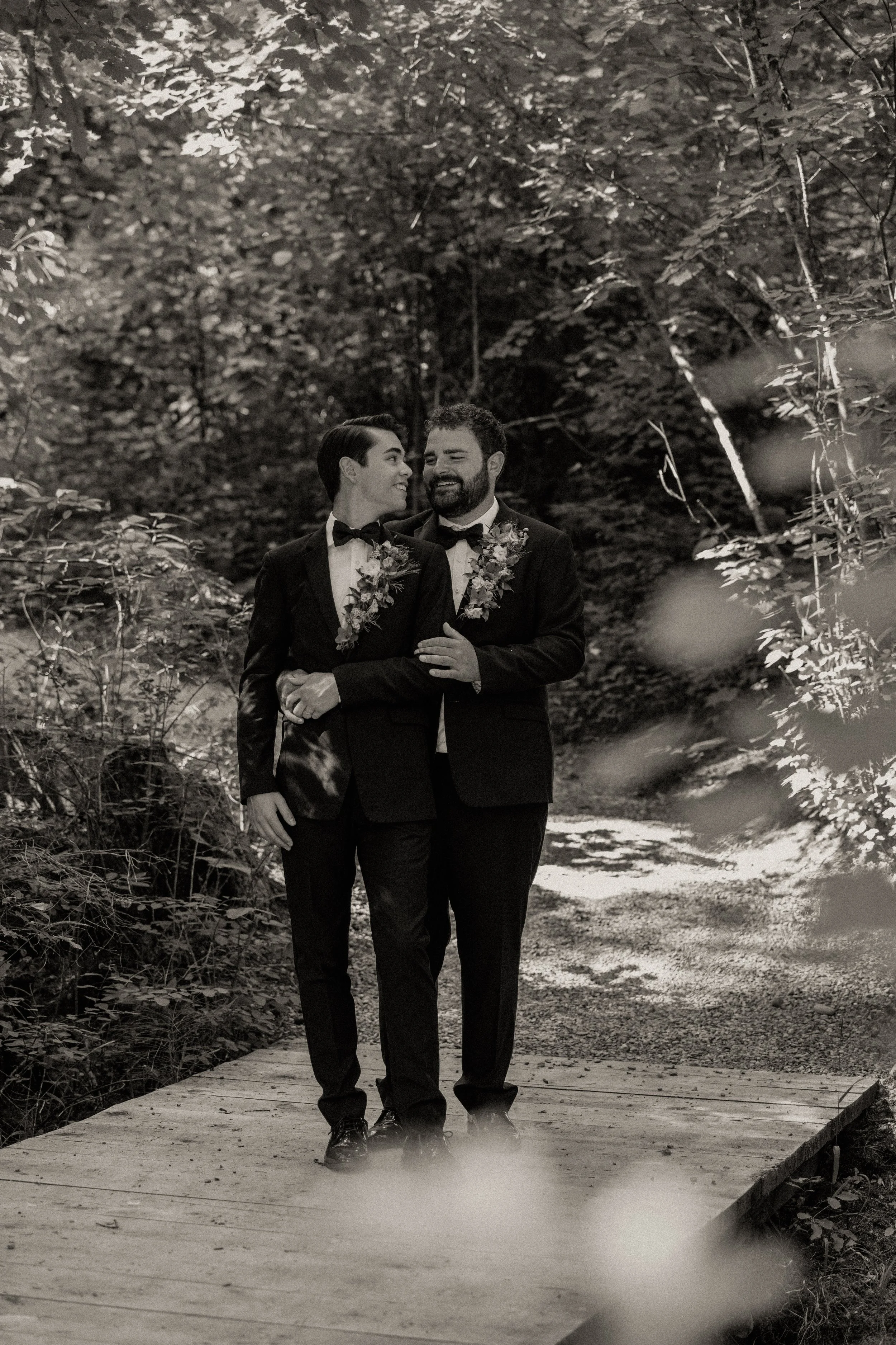 Two men in tuxedos walking arm-in-arm on a wooden bridge in a wooded area, smiling and looking at each other.