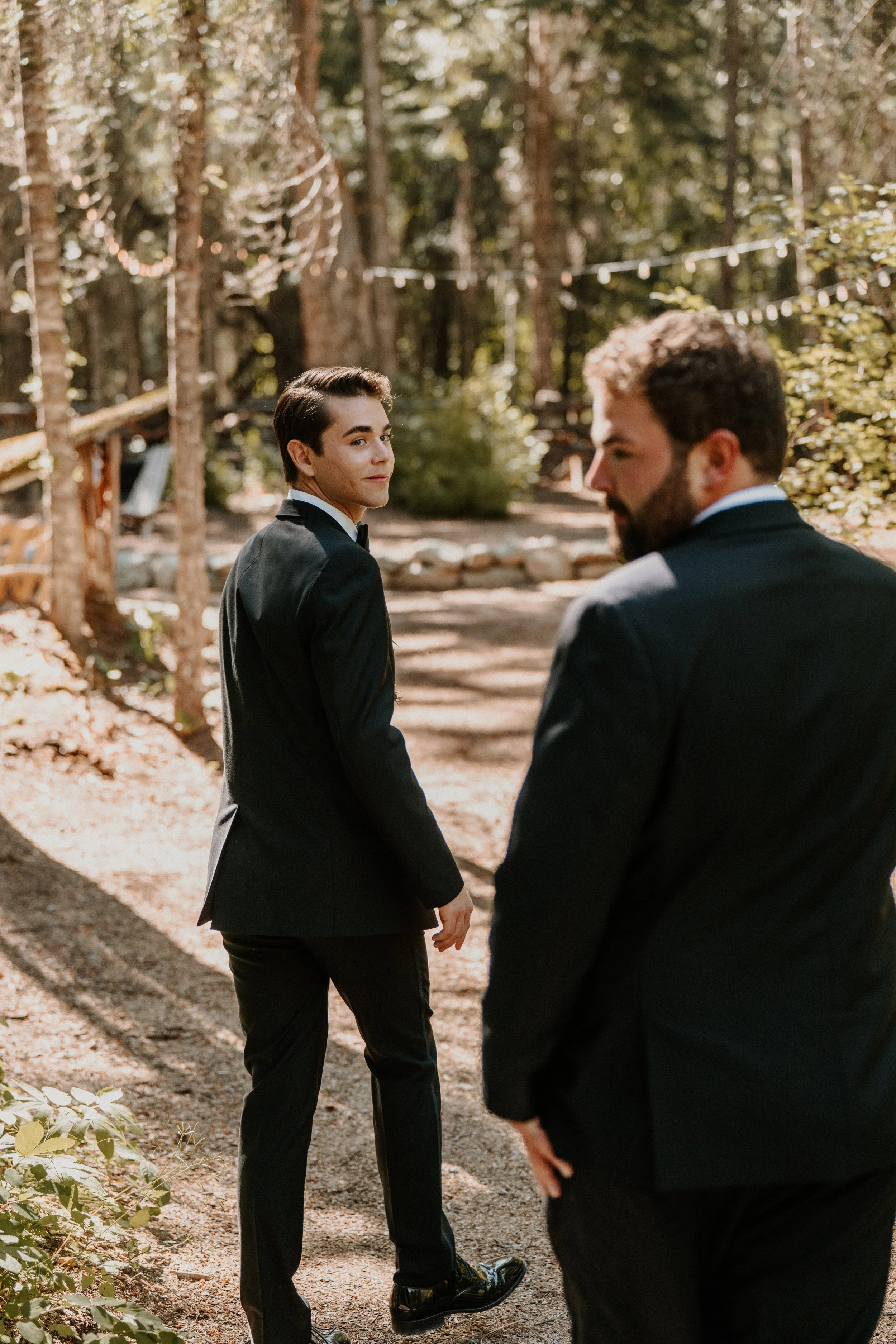 Two men in suits walking on a dirt path through a wooded area with string lights overhead, one looking back at the camera.