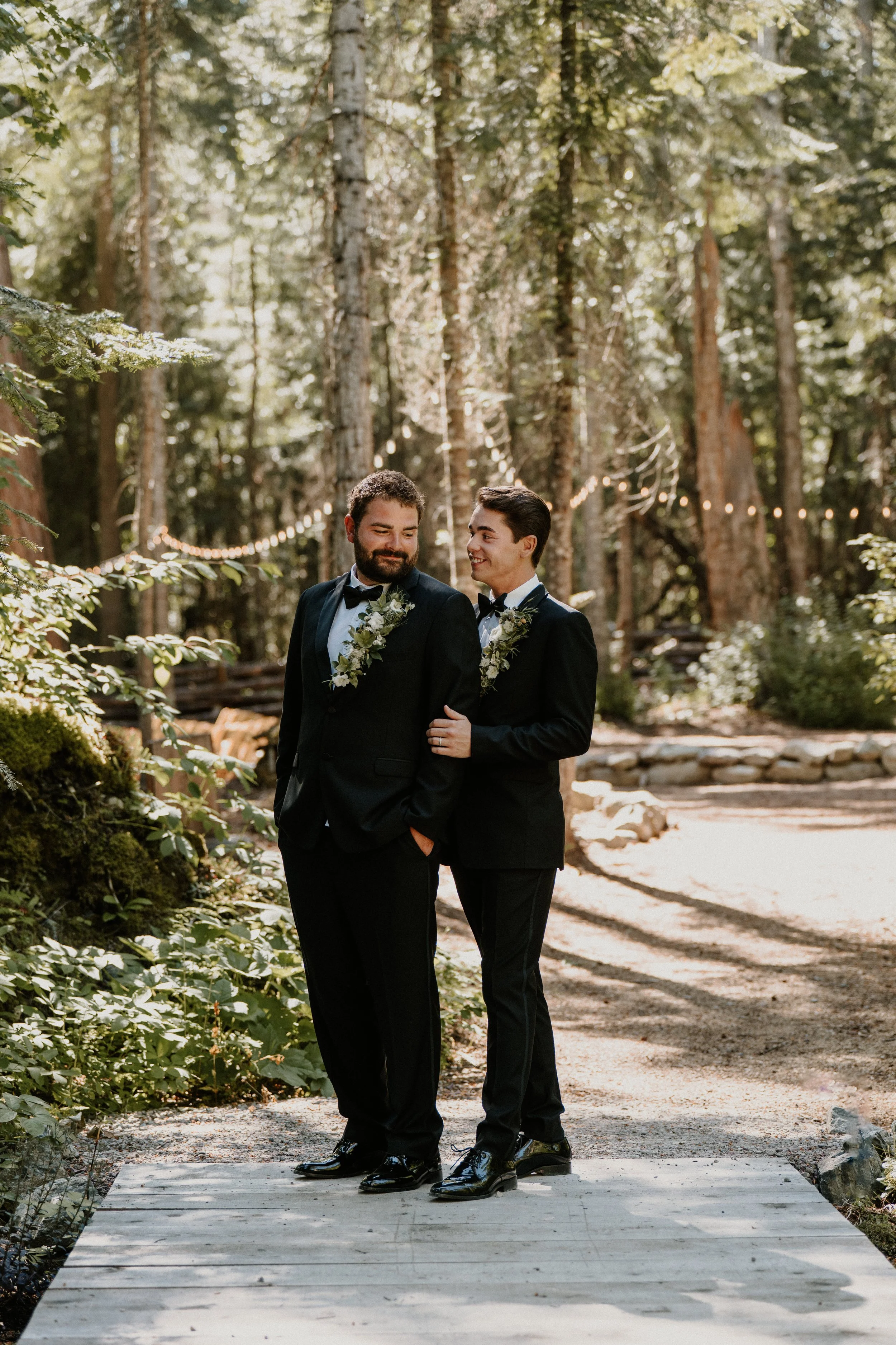 Two men dressed in black tuxedos with floral boutonnières, standing close and smiling at each other, on a small wooden bridge in a forest with string lights in the background.