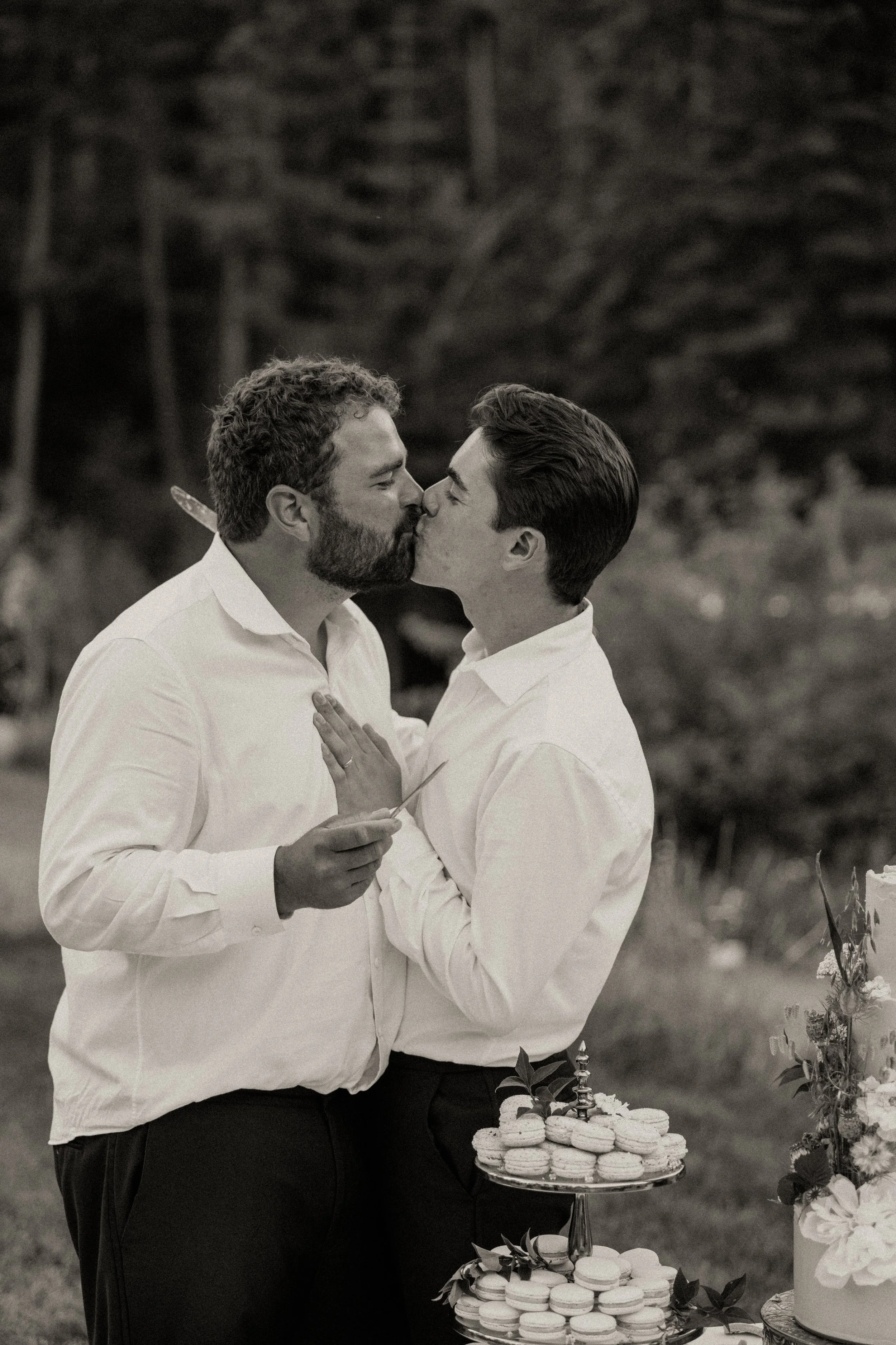 Two men in white shirts kissing at an outdoor celebration, with a table of macarons and a cake in the foreground.