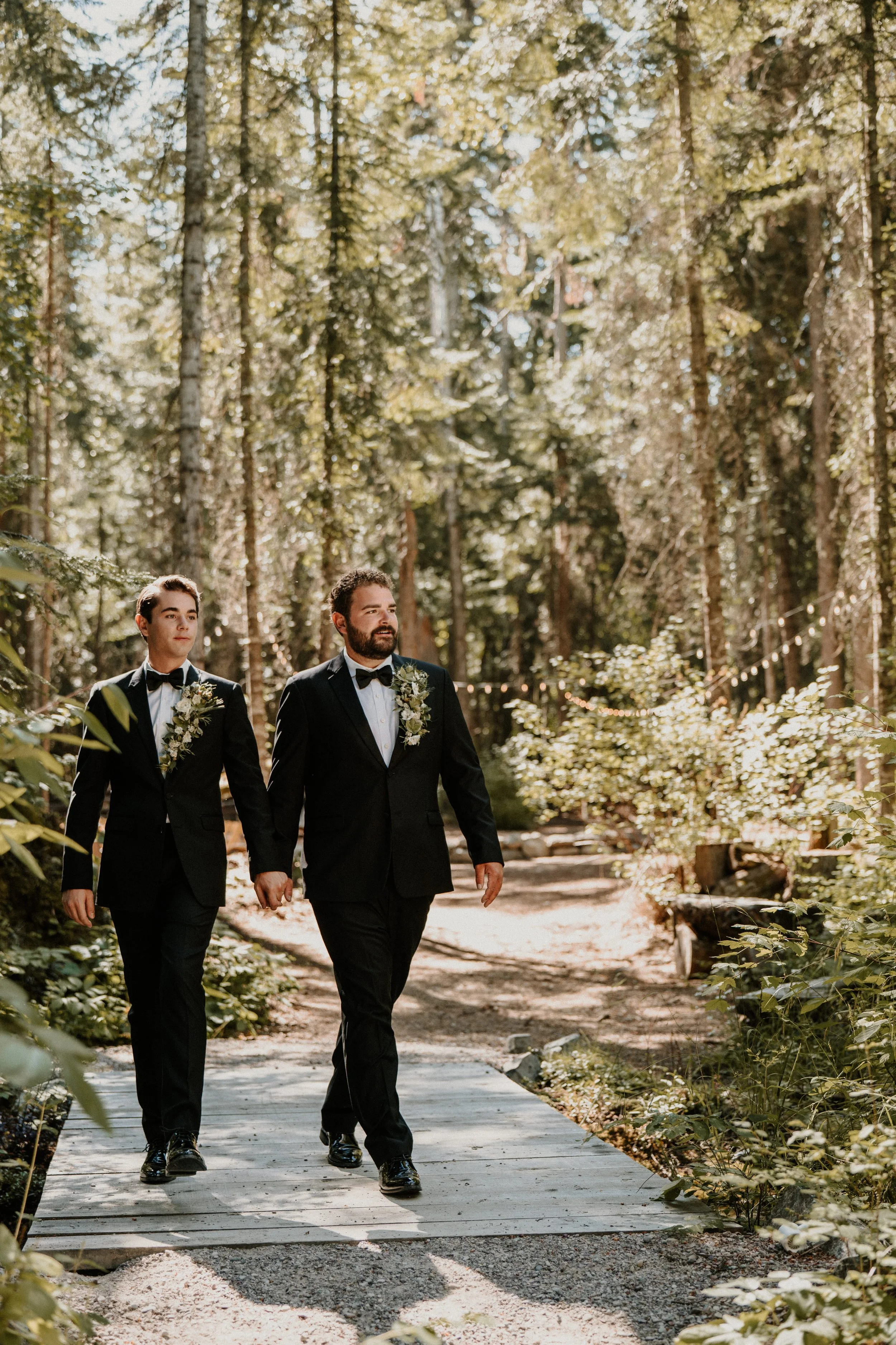 Two men in tuxedos walking hand in hand on a wooded outdoor path, decorated with string lights, during a wedding ceremony.