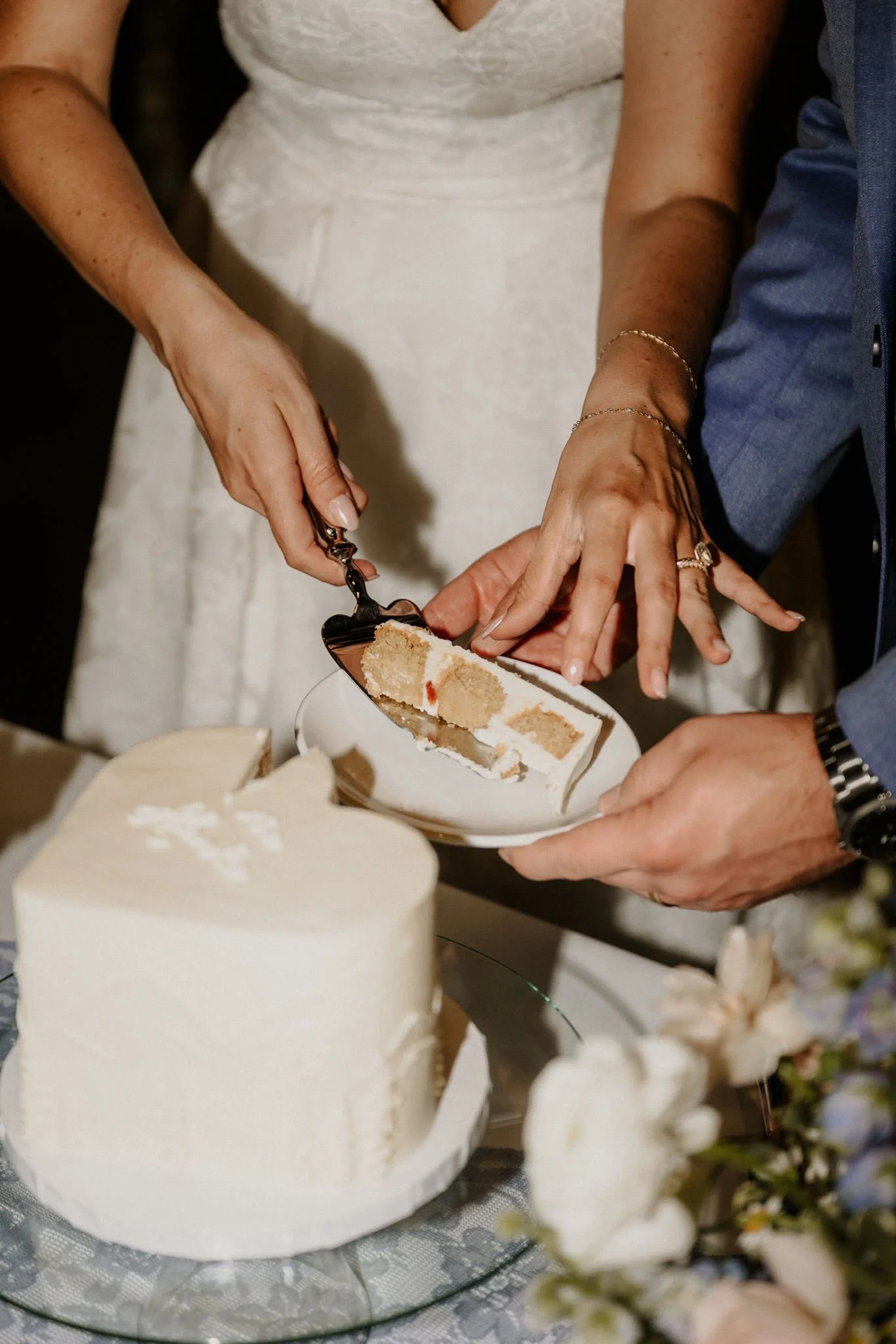 Close-up of a person cutting a white frosted wedding cake with a knife. The person is wearing rings and bracelets. The cake is on a glass platter, and there are flowers in the foreground.