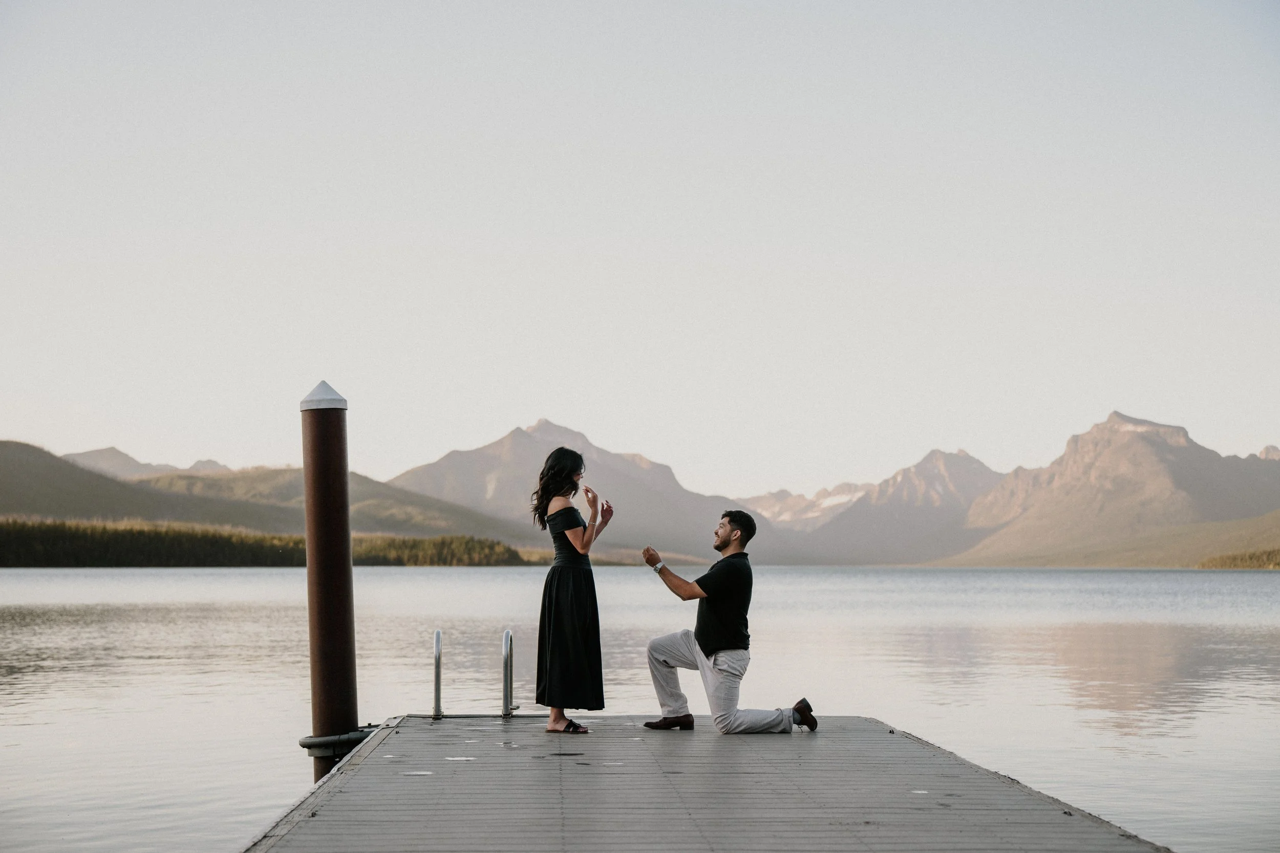 A man proposing marriage to a woman on a dock by a lake in Glacier National Park, with mountains in the background