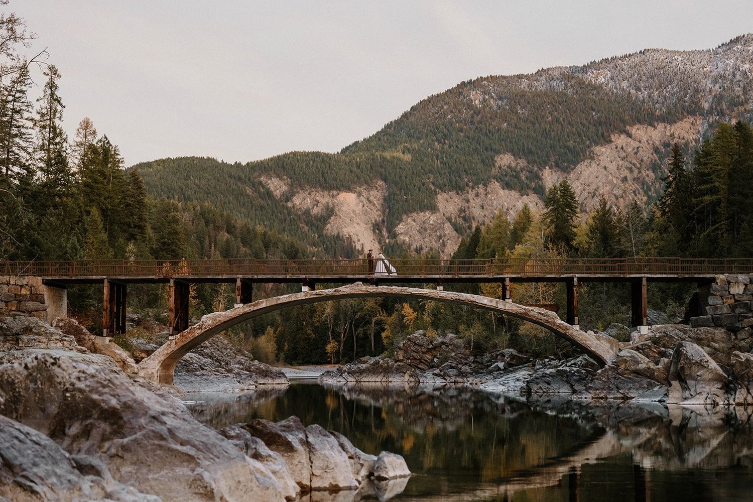 Couple on a wooden bridge over a rocky river with mountains behind