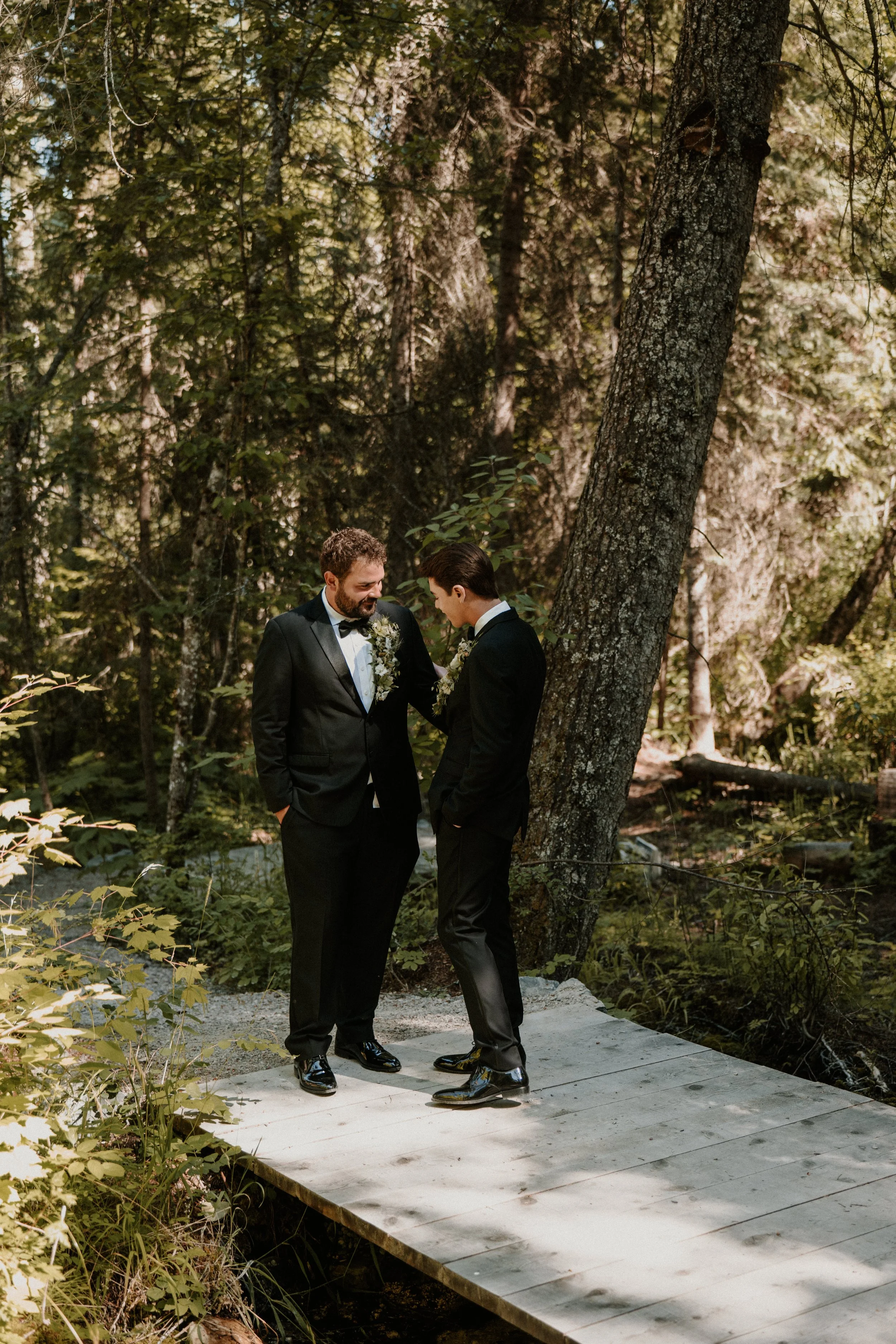 Two men in tuxedos standing on a wooden bridge in a forest, engaging in a heartfelt moment with one touching the other's chest, surrounded by trees and greenery.