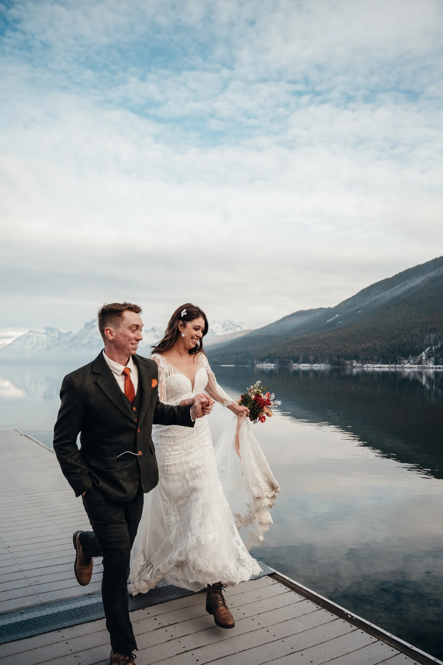 Bride and groom walking the Apgar dock together with snowy peaks and a cloudy winter sky behind them