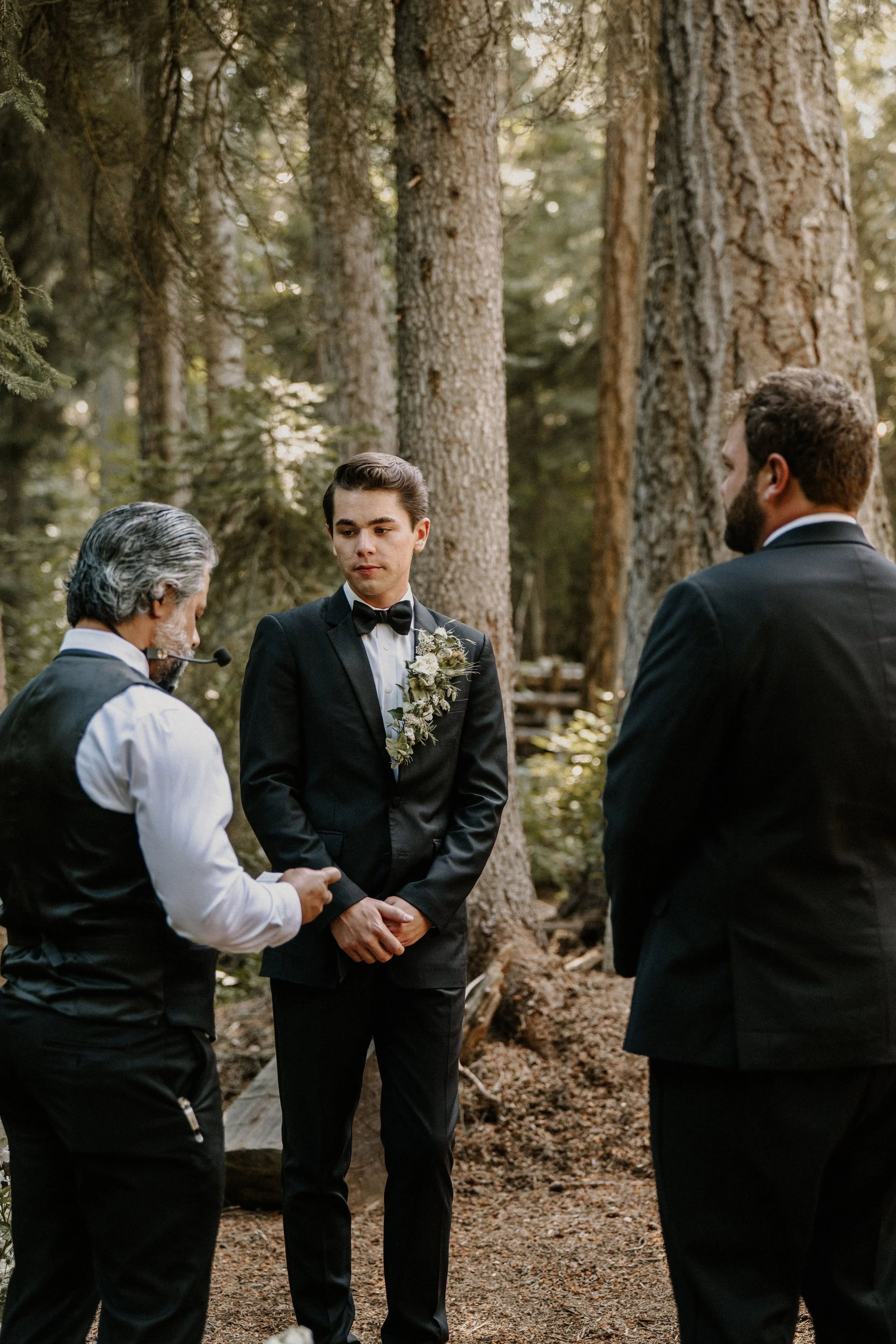 A wedding ceremony taking place in a forest, with a groom standing at the altar and two men in suits, one officiating, during daytime.