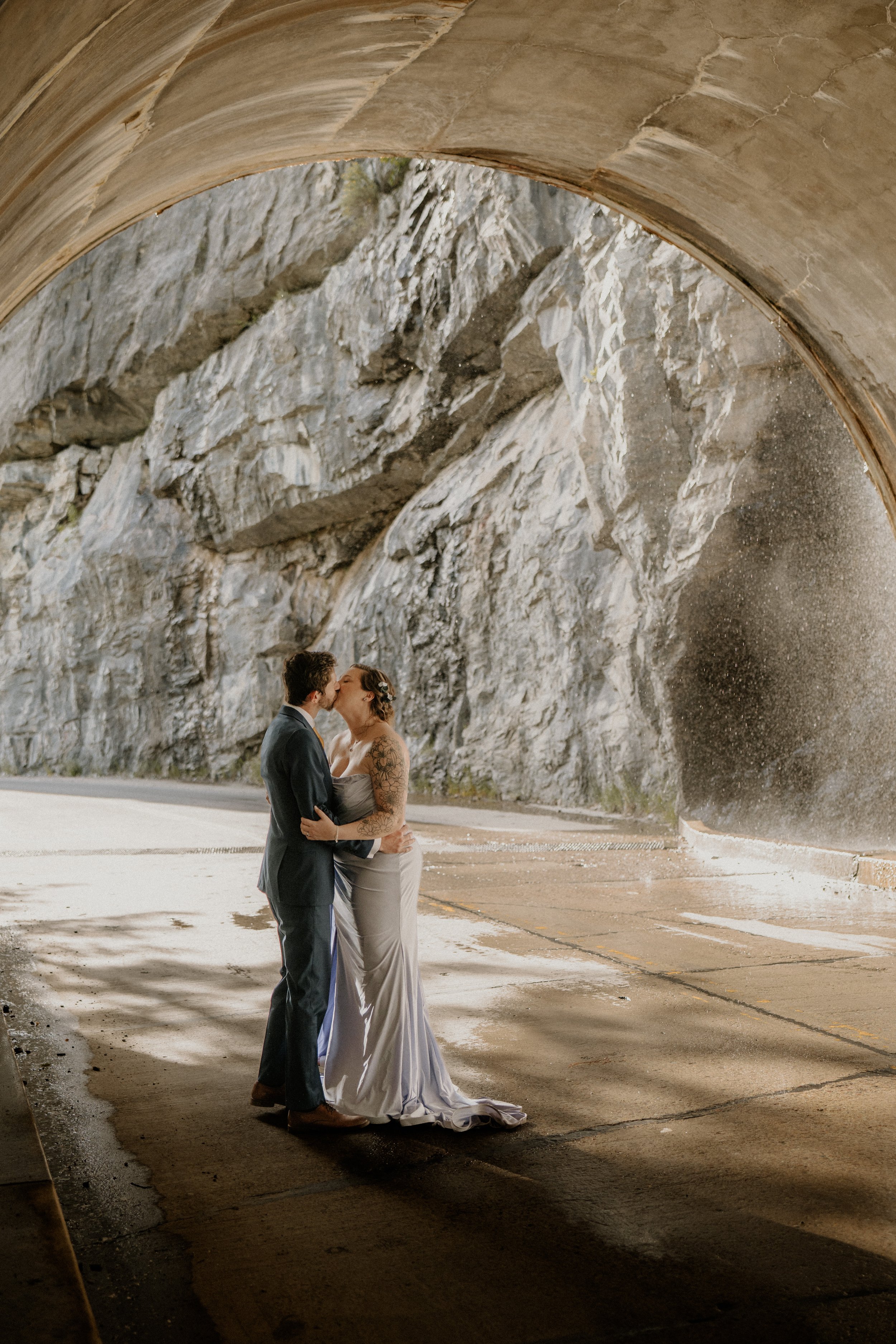 A couple dressed in wedding attire sharing a kiss beneath a large rocky tunnel with an arched concrete ceiling.