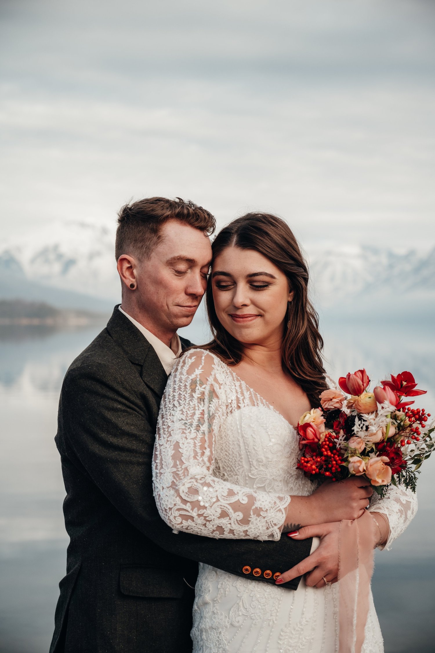 Bride holding her bouquet while embracing the groom by Lake McDonald with snow-covered mountains behind them