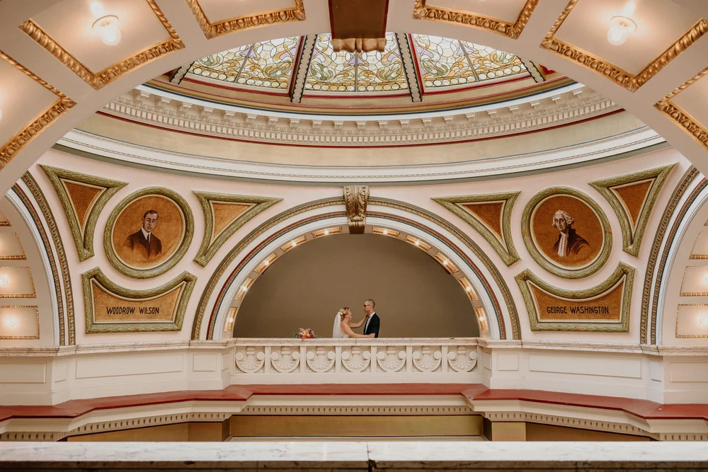 Couple on a balcony inside a grand historic building with a stained glass ceiling