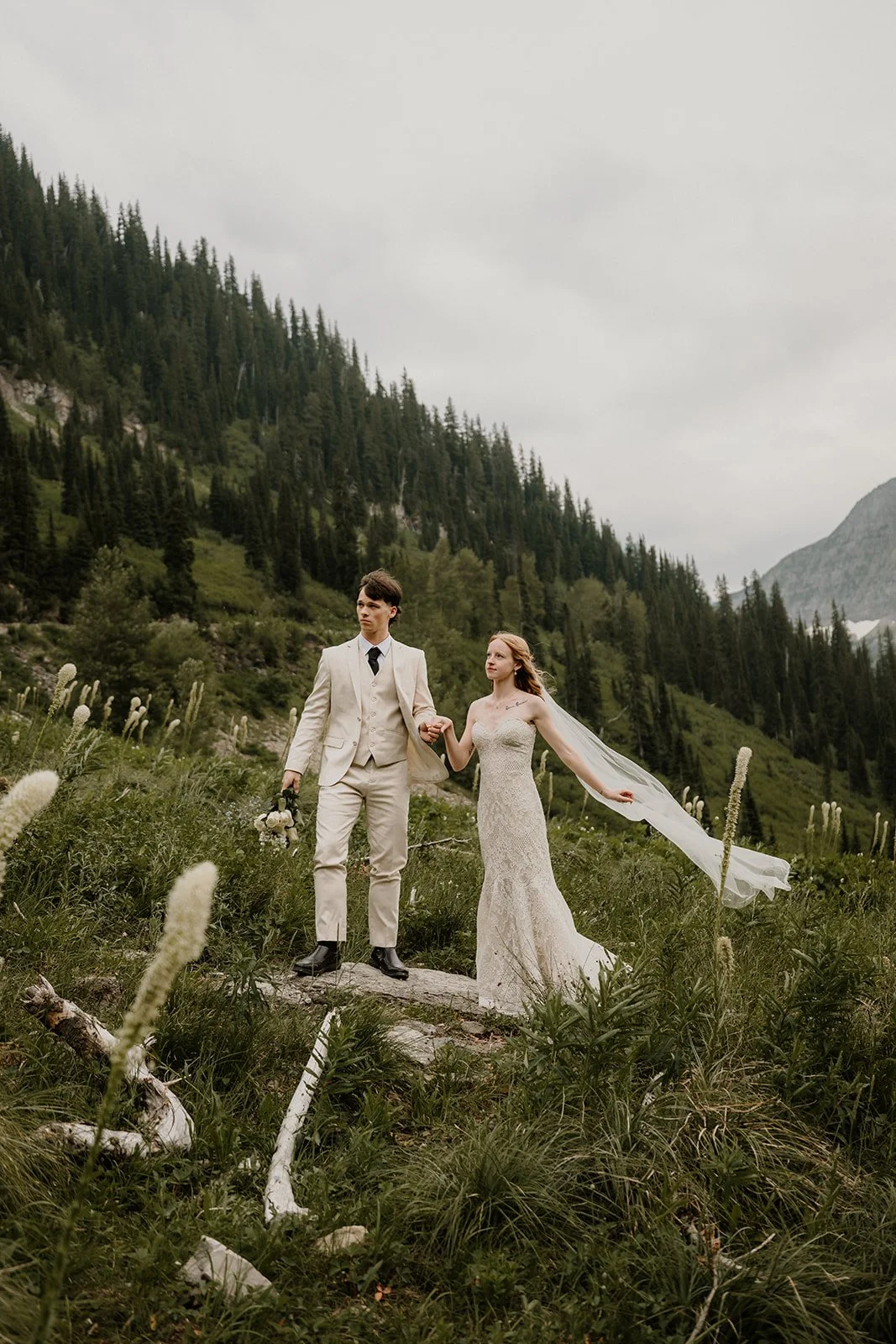 A bride and groom walking hand in hand in a lush, mountainous outdoor setting, with the groom wearing a cream-colored suit and the bride in a long white lace wedding dress and veil.