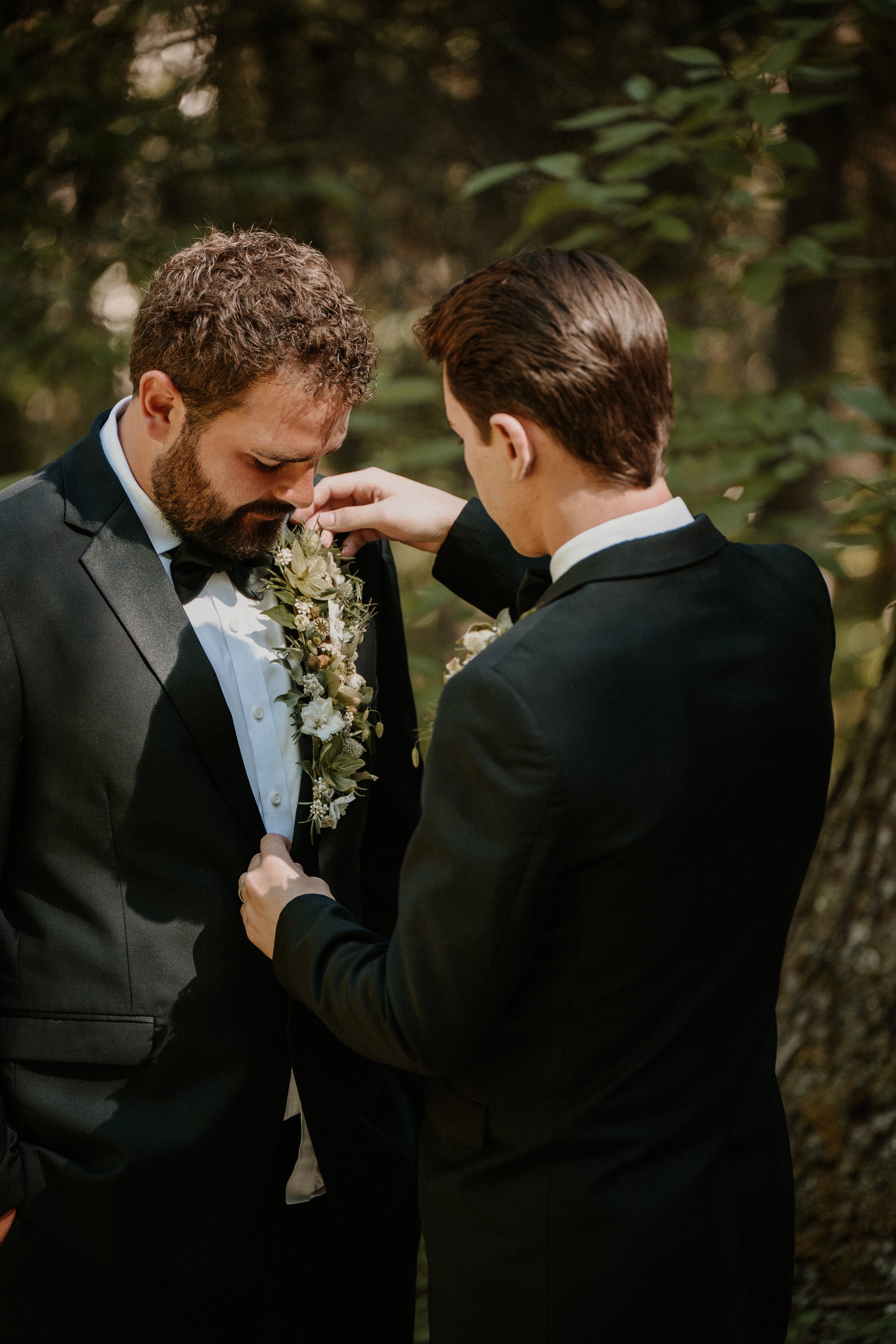 Two men in black tuxedos sharing an intimate moment outdoors, with one placing a boutonniere on the other's lapel during a wedding or formal event.