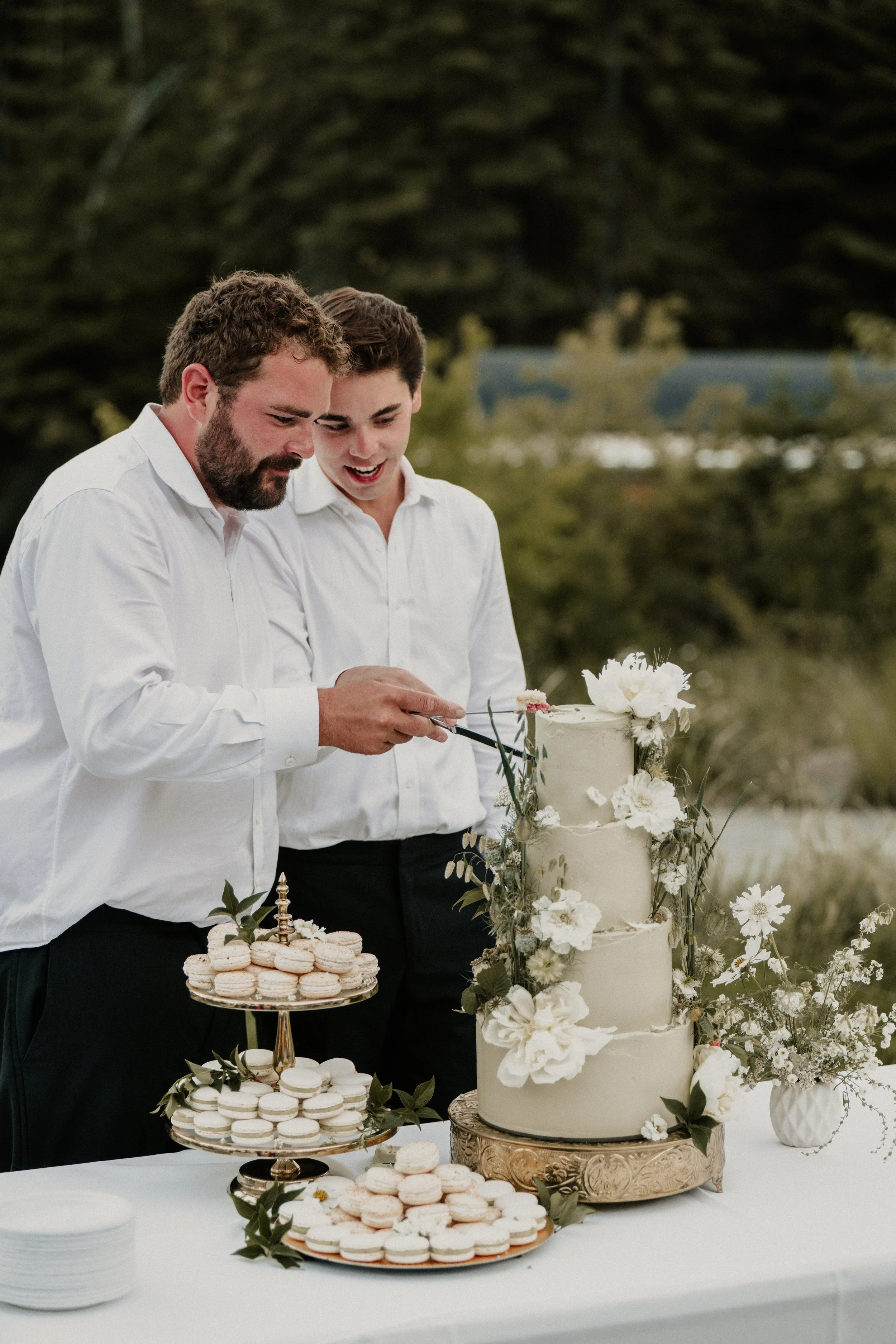 Two men in white shirts cutting a wedding cake outdoors, with macarons on plates nearby and a floral arrangement on the table.