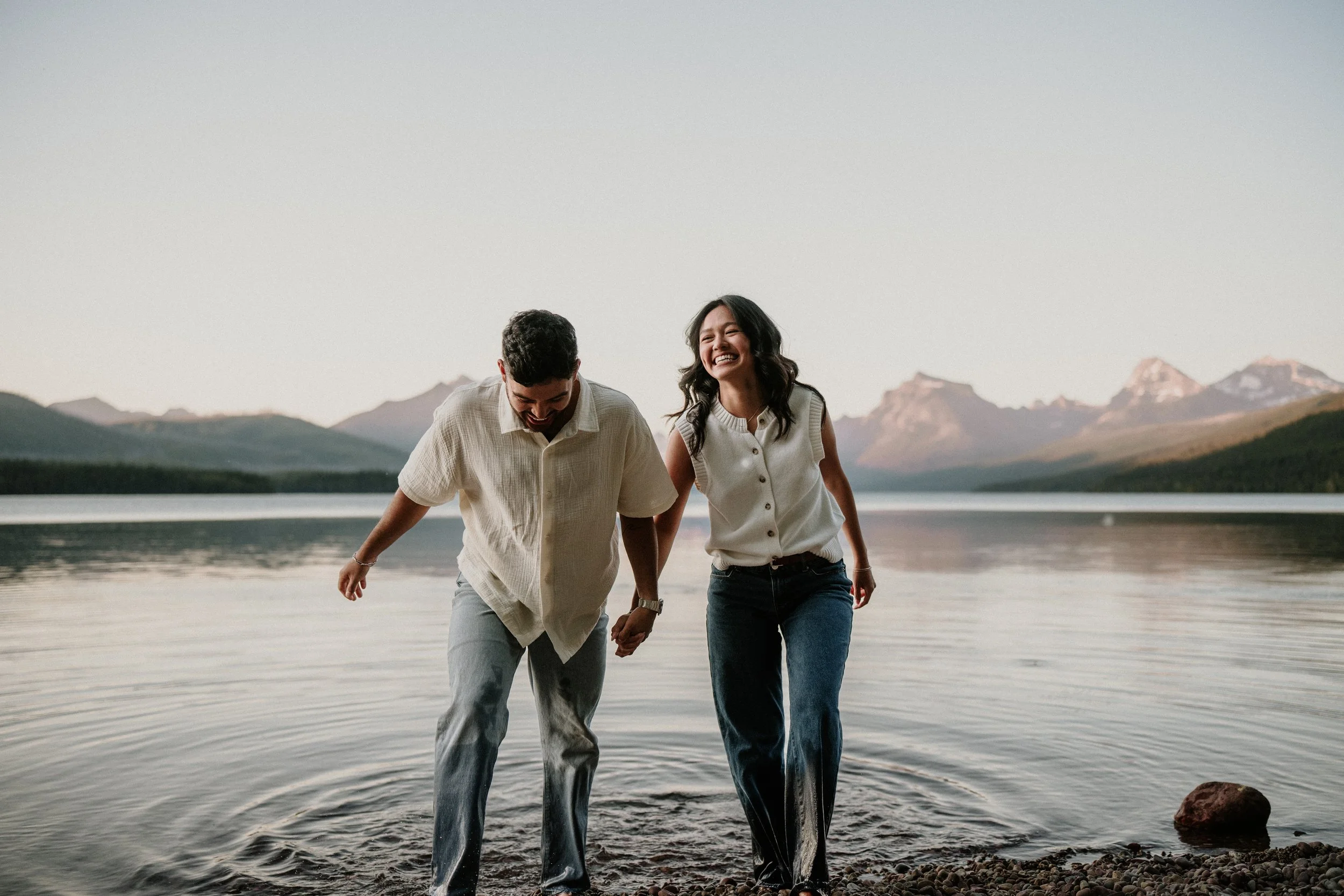 Newly engaged couple holding hands and splashing into a Glacier National Park lake with mountains behind