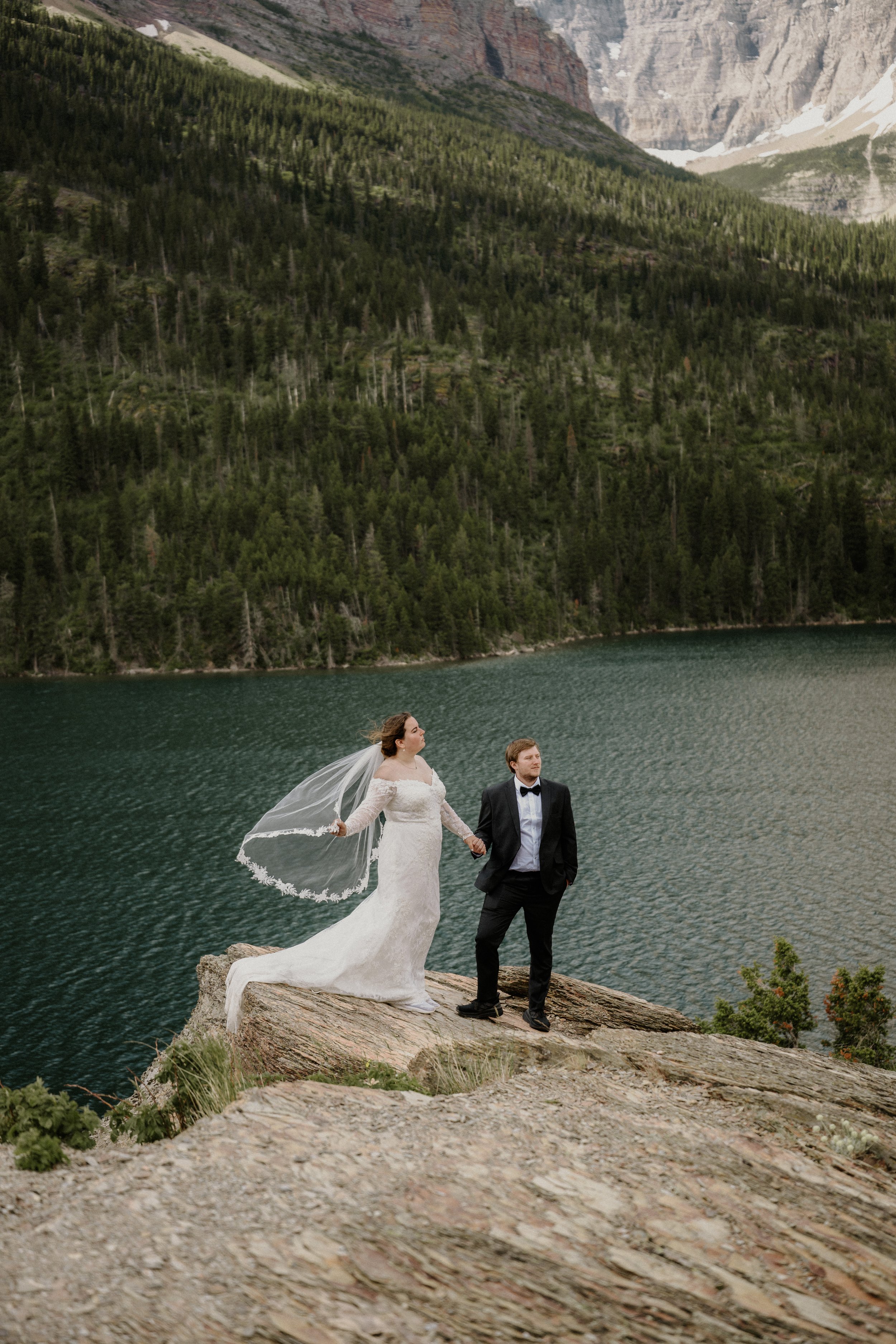 A bride and groom standing on a rocky outcrop near a lake with mountains and dense forest in the background.