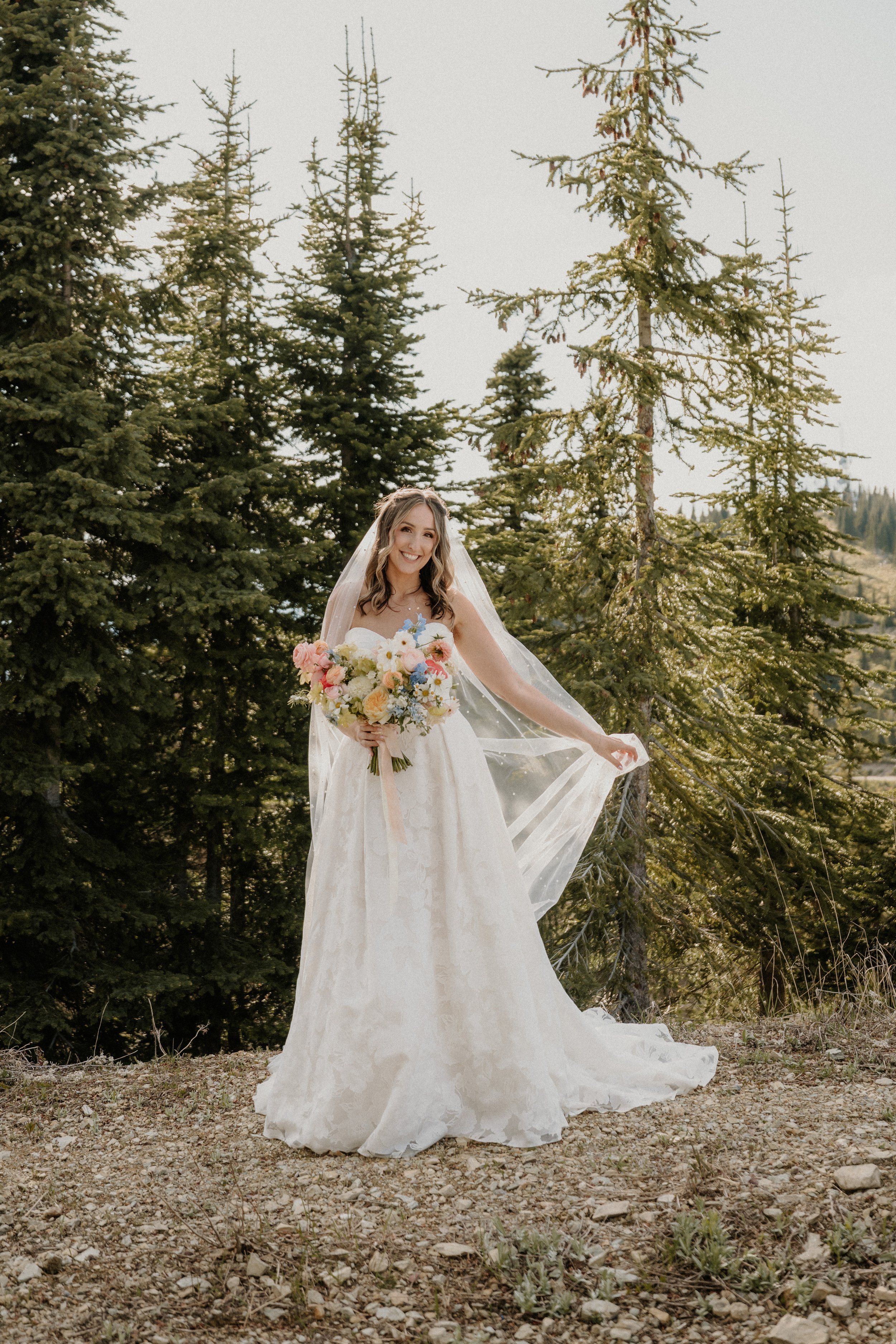 A woman in a white wedding dress holding a bouquet of flowers, standing outdoors among tall evergreen trees.