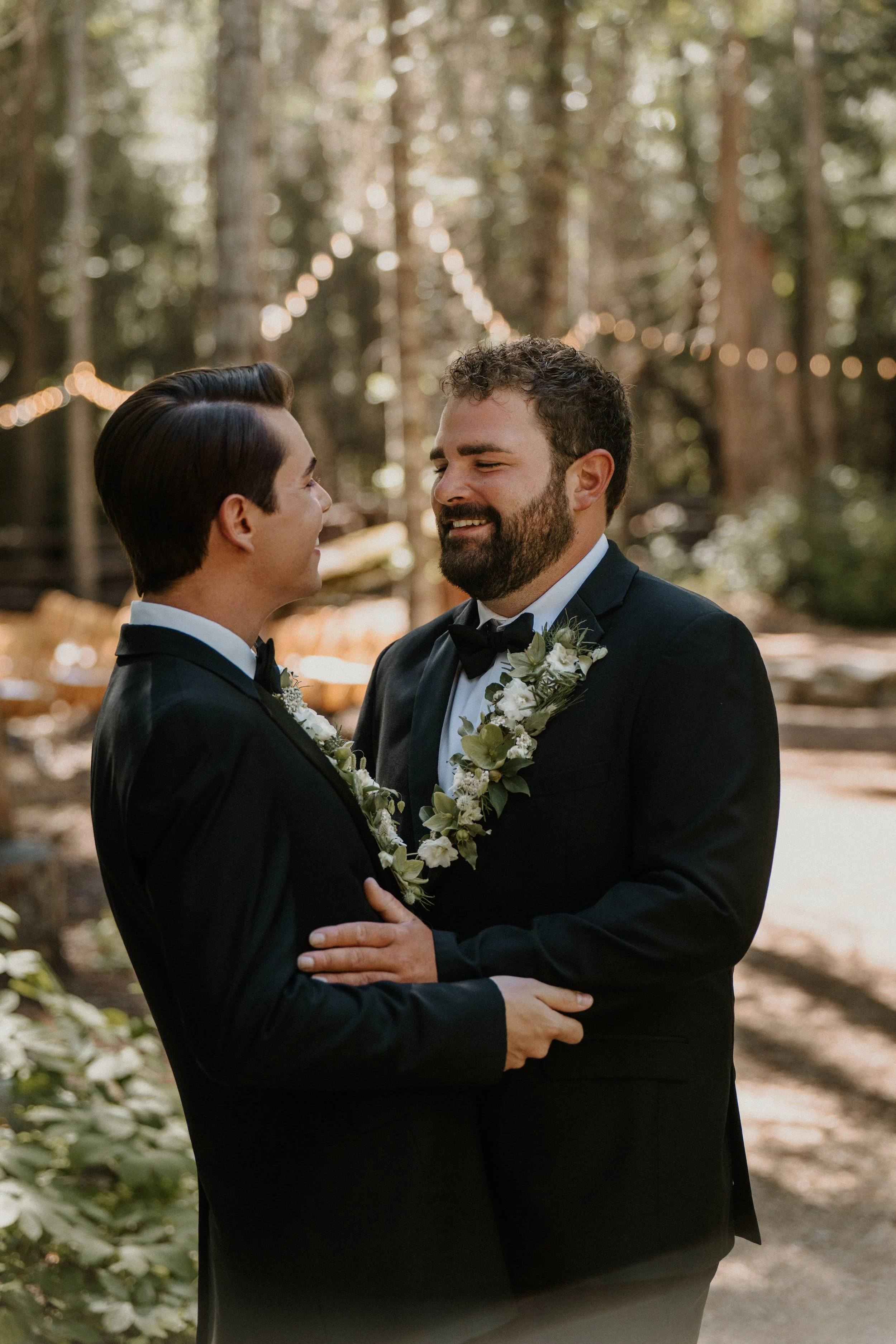 A couple dressed in black tuxedos and floral leis face each other in a forest, smiling, with string lights in the background.