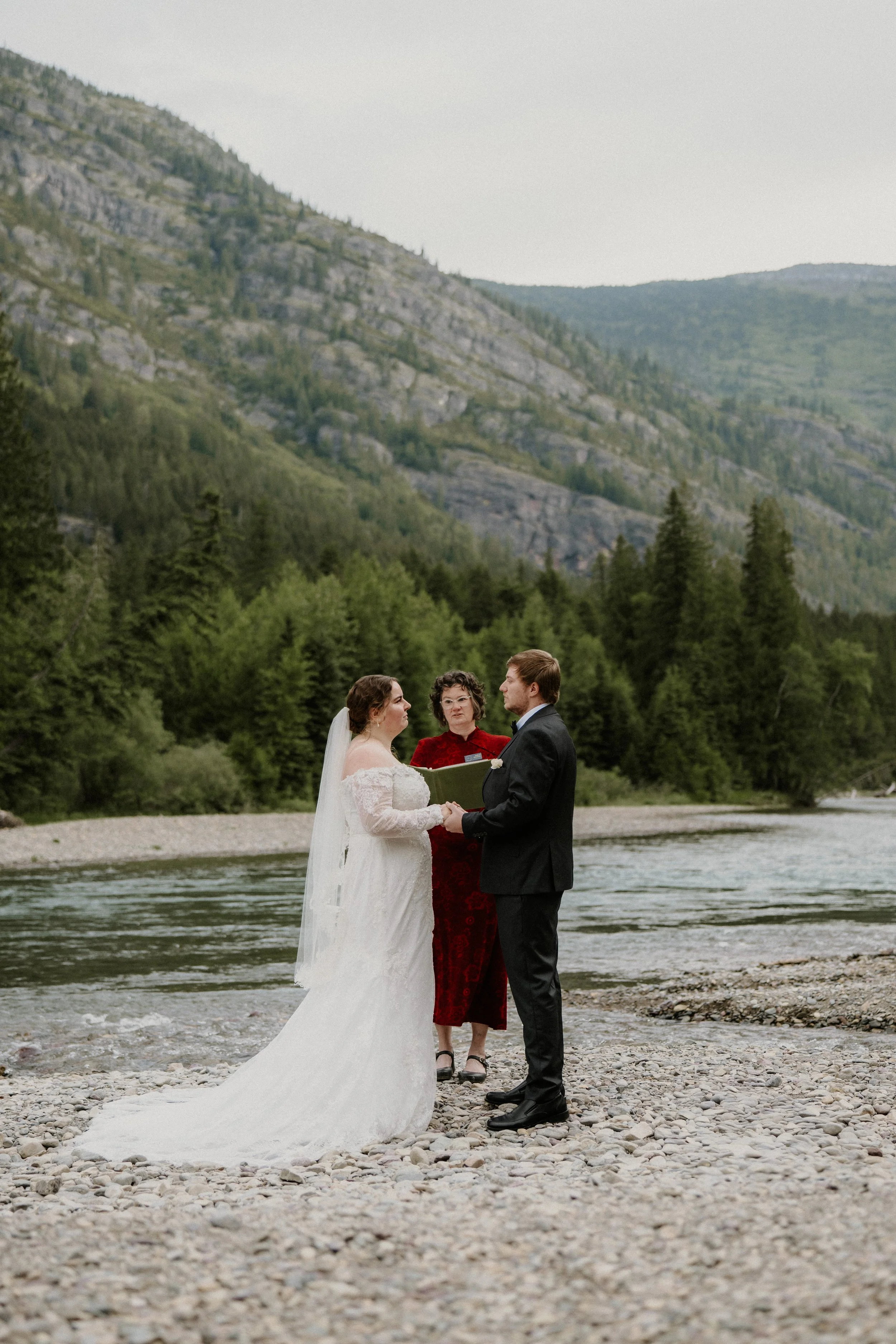 A bride and groom standing in front of a river exchanging vows during a wedding ceremony, with a river, trees, and mountains in the background.