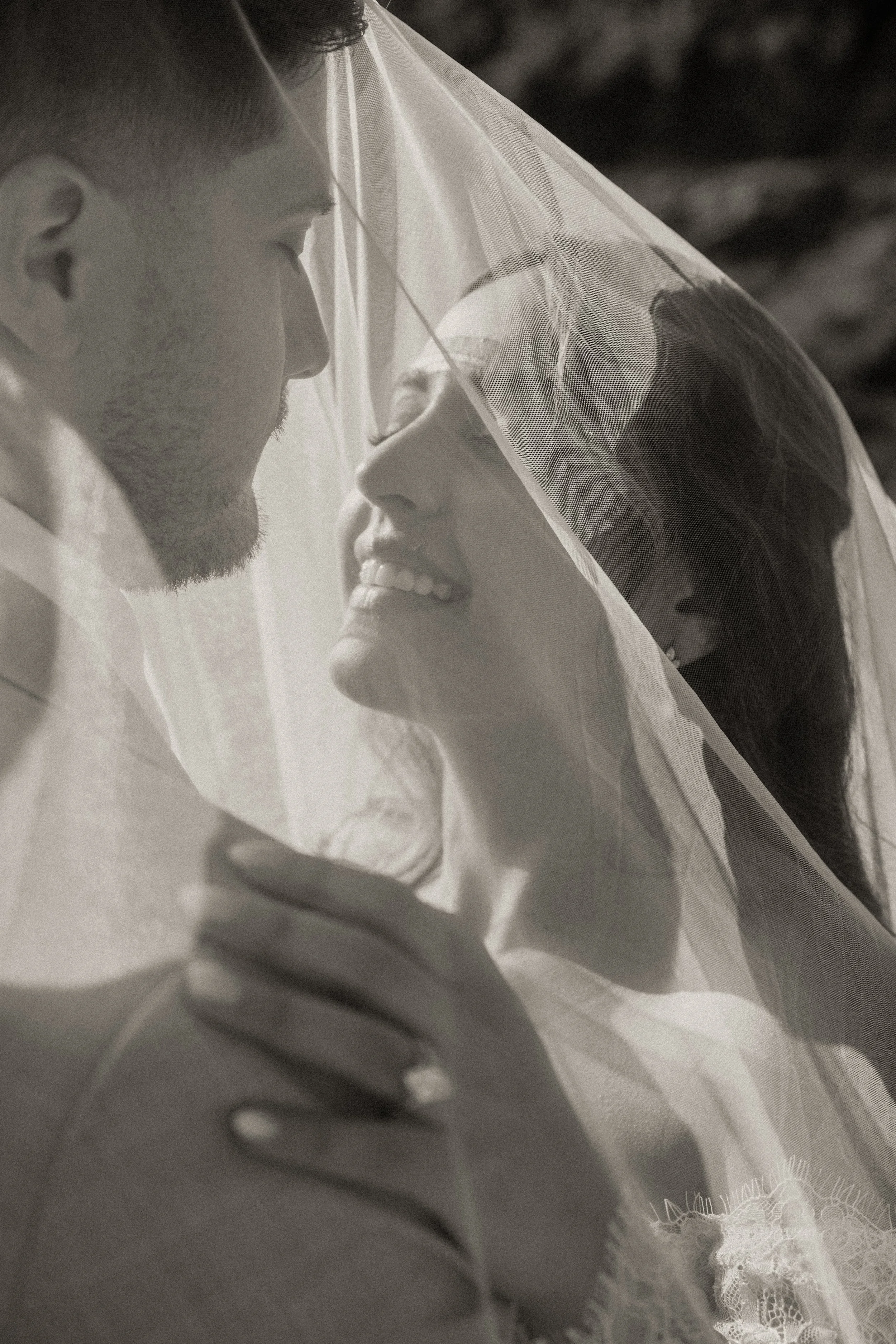 A man and woman close together, with the woman smiling and the man appearing to gently touch her face, under a veil with lace details.