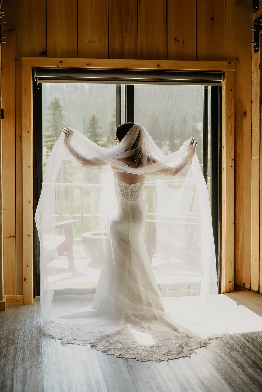 A bride in a white wedding dress holding her veil, standing inside in front of a large glass door that looks out to a porch and a wooded landscape beyond.