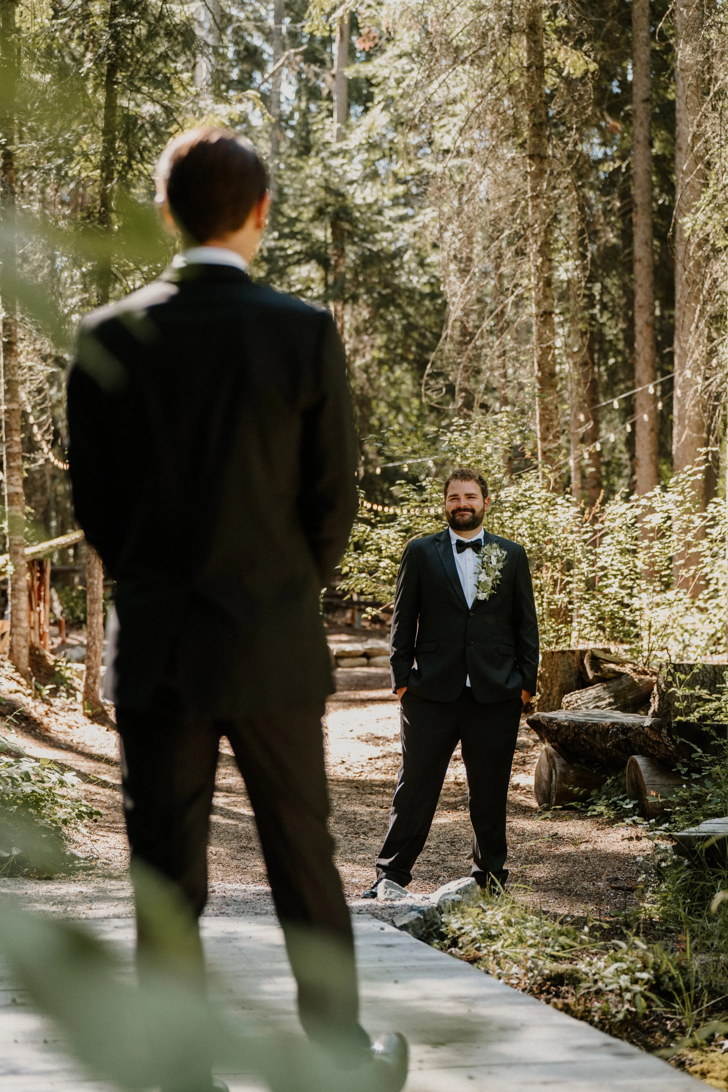 A person in a black suit, standing with his hands in his pockets, faces another person also in a black suit on a forest trail. The person facing away has short dark hair, and the person facing forward has a beard and a floral boutonniere. Trees and s