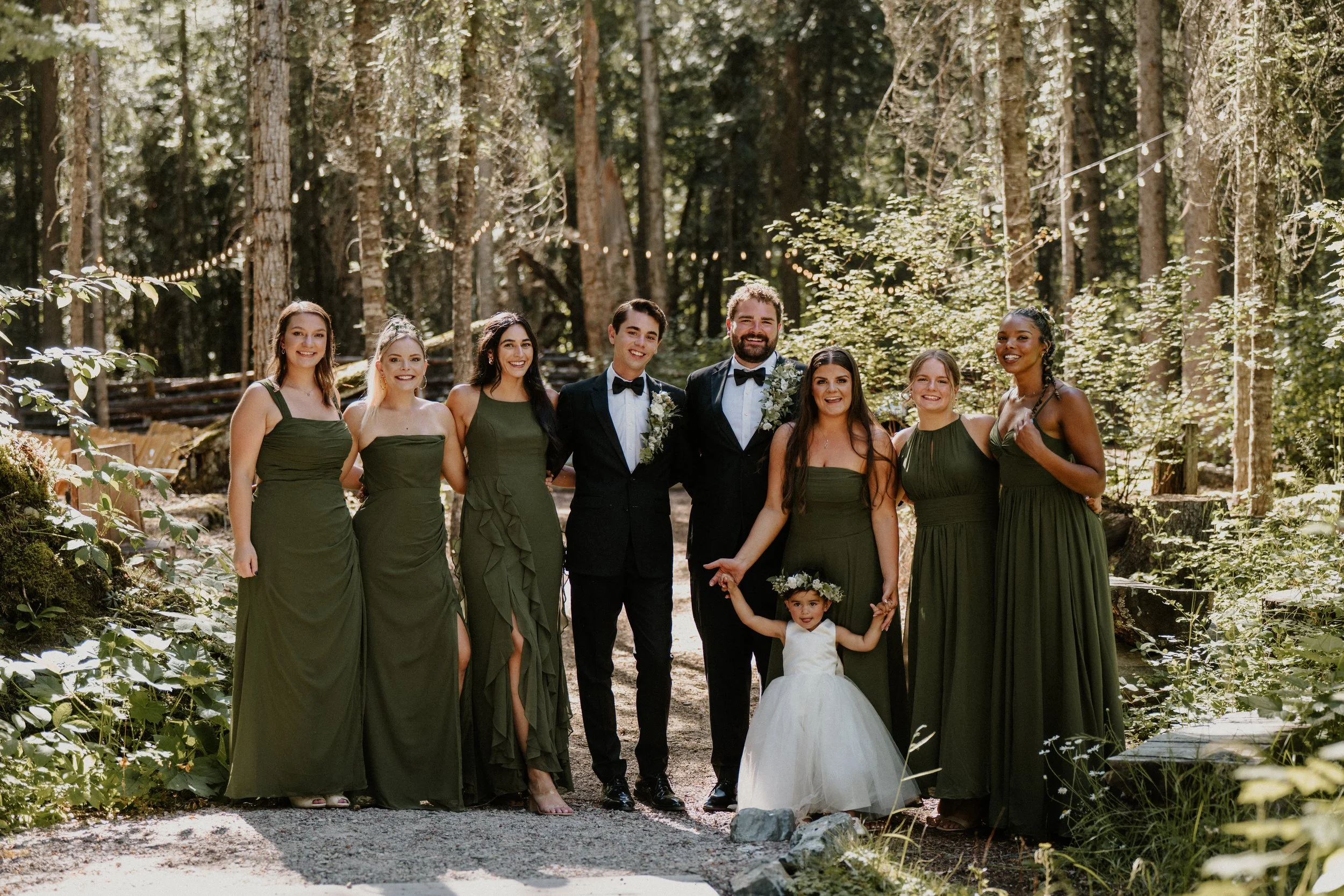 A wedding party standing in a lush, green forest with sunlight filtering through the trees. The group includes women in matching green dresses, two men in black tuxedos, a young girl in a white dress and floral crown, and a woman in a green dress and