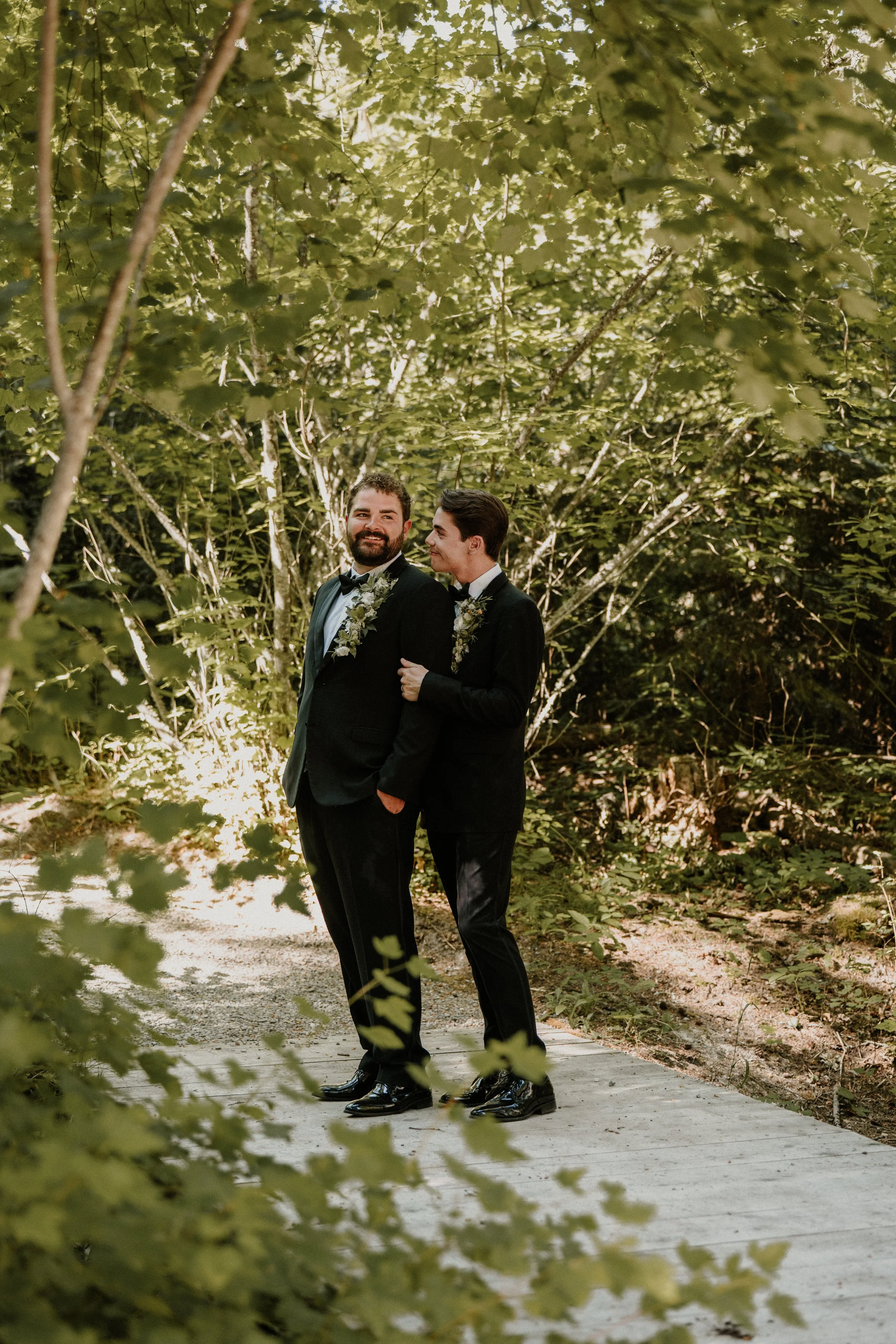 Two men in tuxedos with floral garlands on their necks, standing on a pathway surrounded by lush green trees.