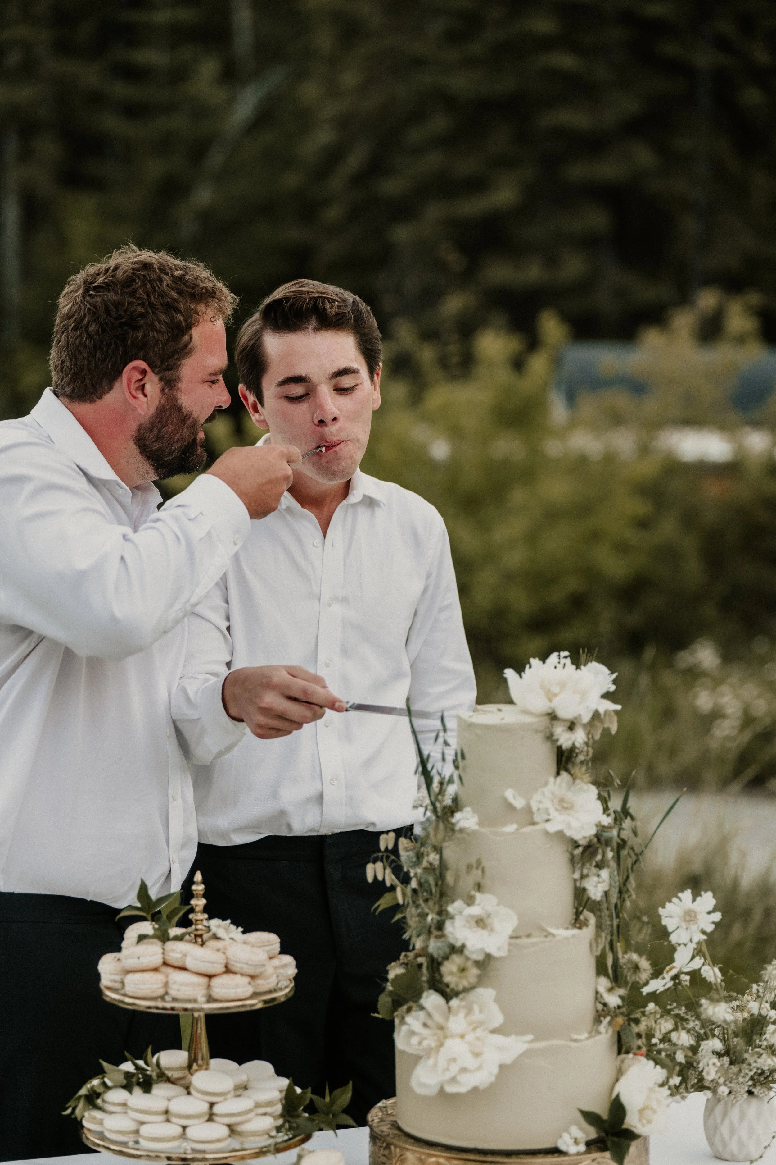 Two men in white shirts cutting a wedding cake outdoors, with a three-tiered white cake decorated with white flowers and greenery, accompanied by macarons on a tiered stand.