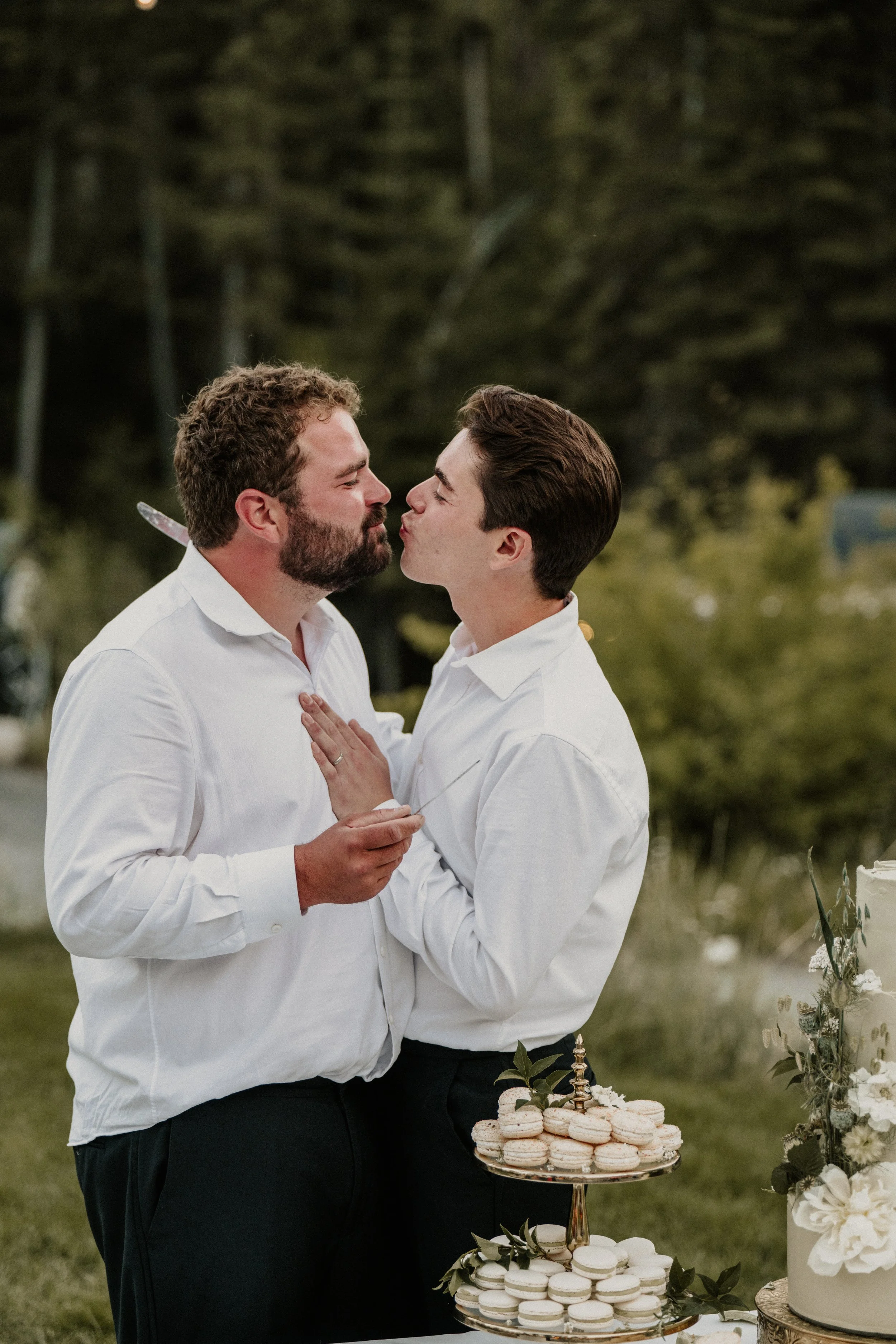 Two men dressed in white shirts sharing a kiss at an outdoor wedding reception with a cake and macarons displayed on a tiered stand nearby.
