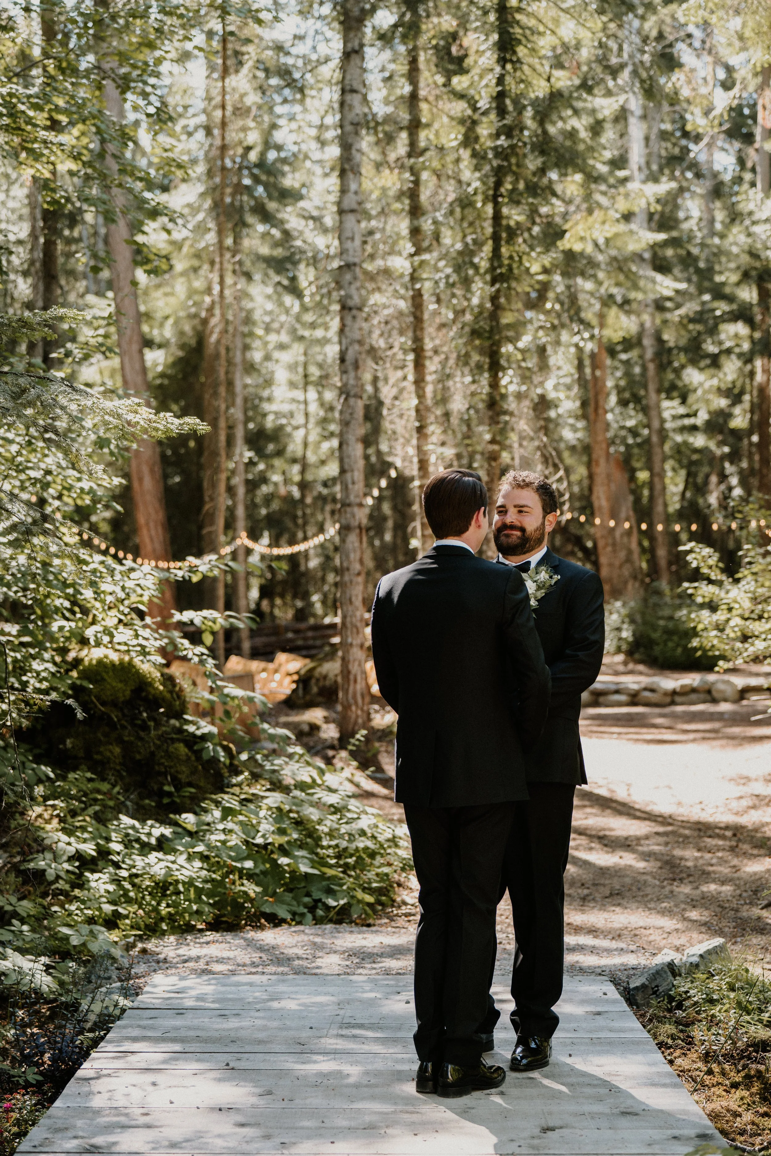 Two men in suits standing and facing each other on a wooden path in a forest, smiling. There are string lights draped between trees in the background.