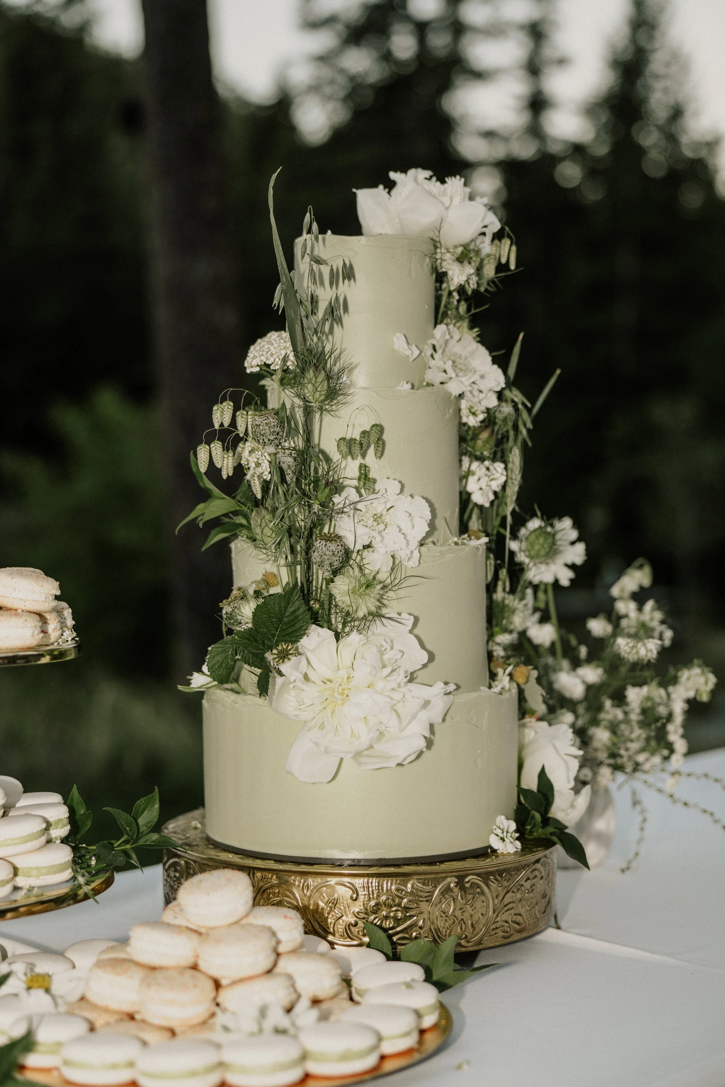 Three-tiered wedding cake decorated with white flowers and greenery, placed on an ornate gold cake stand. Surrounding the cake are plates of macarons and cookies. The setting is outdoors with trees in the background.