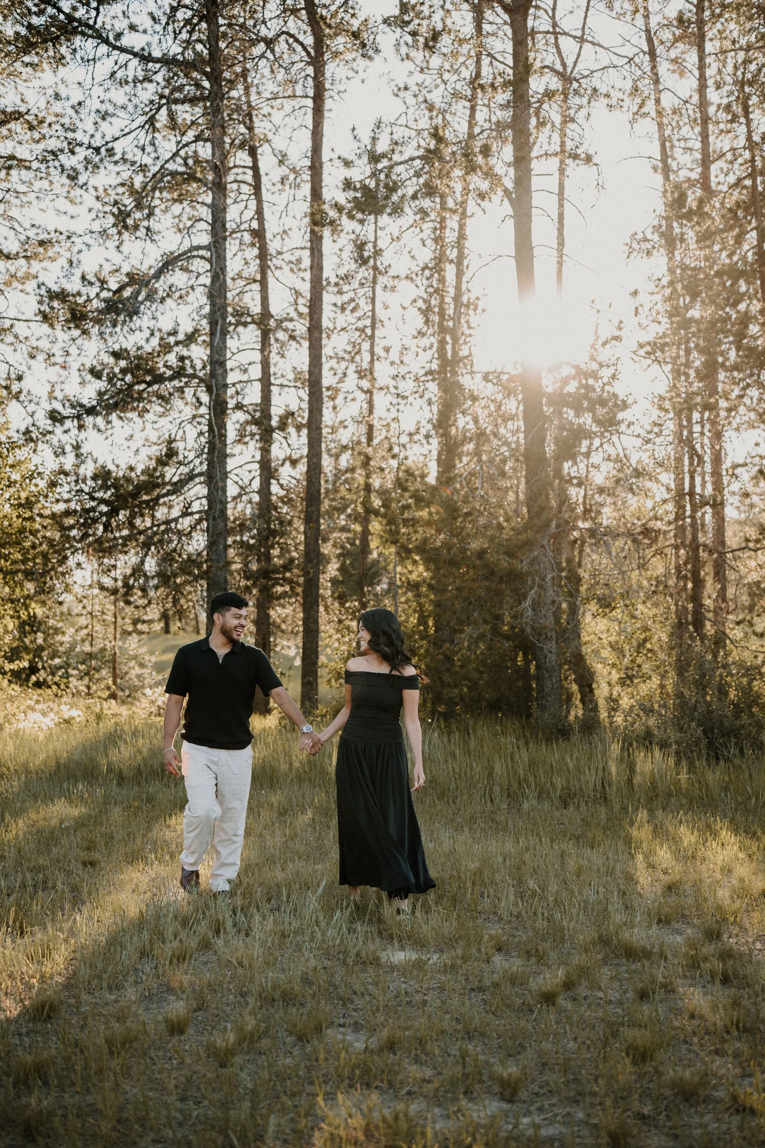 Couple holding hands walking through a grassy field with sun streaming through the trees in Glacier