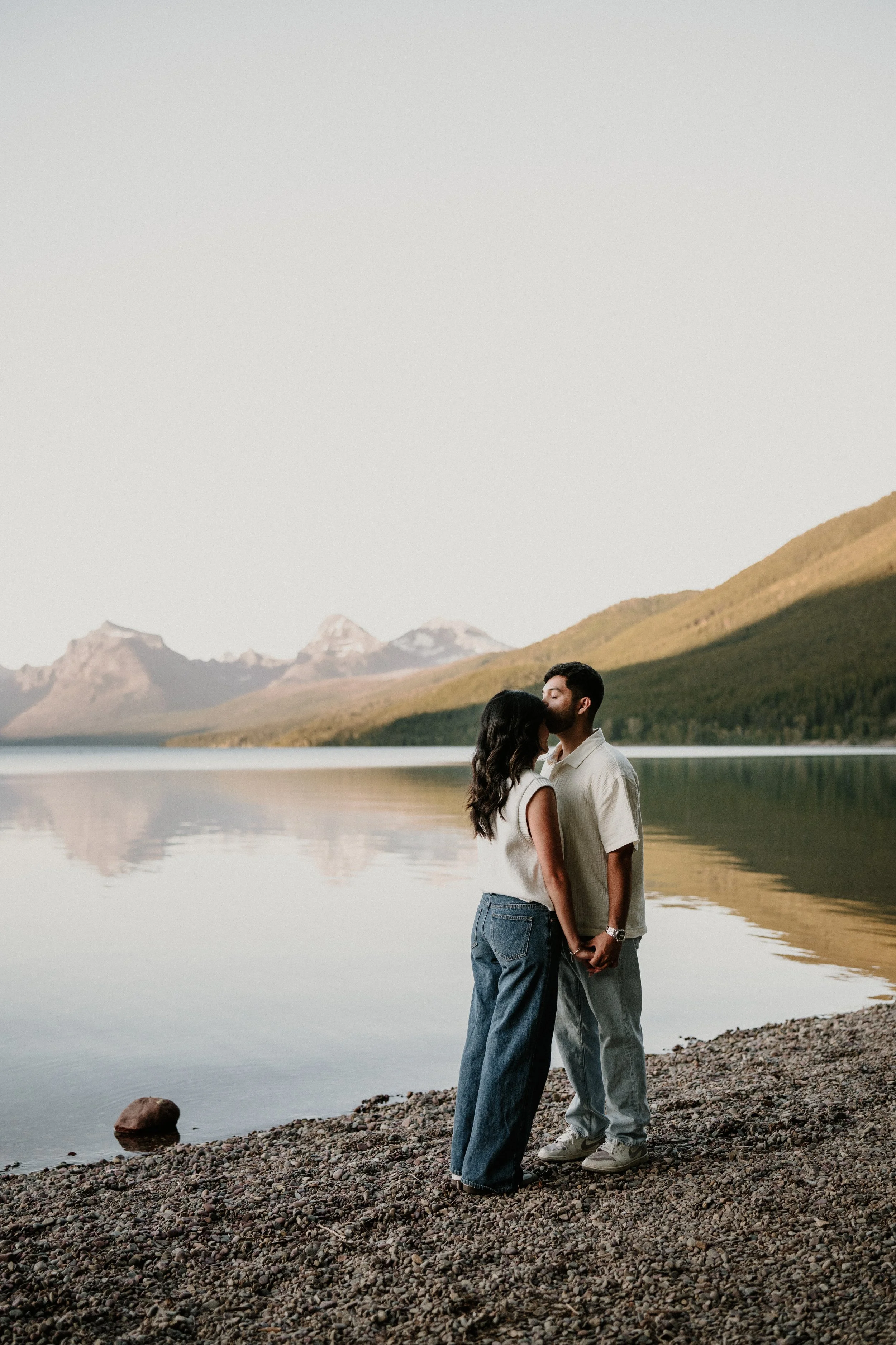 Couple about to kiss on a rocky Glacier National Park lakeshore with mountains behind