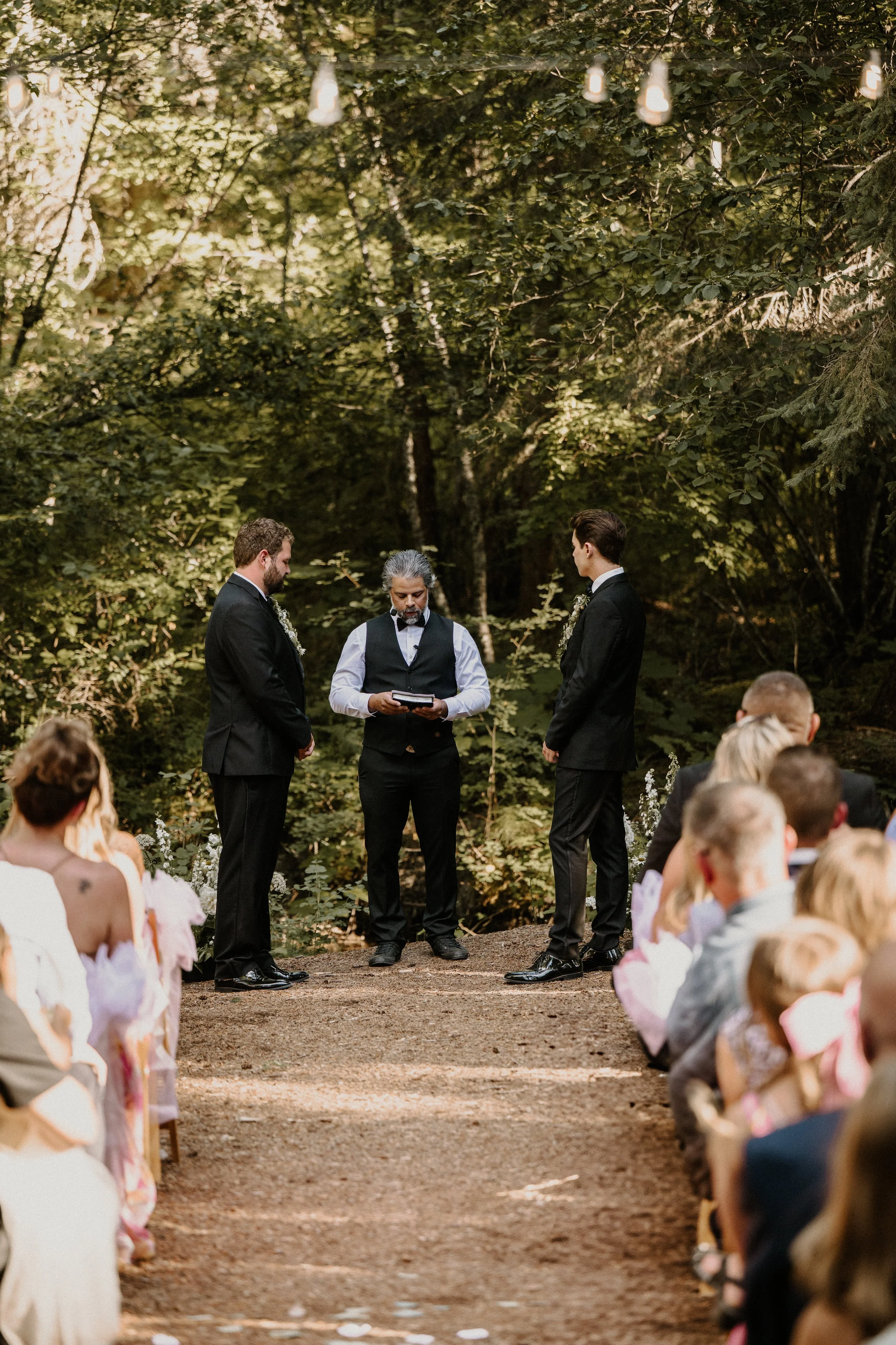 A wedding ceremony taking place outdoors in a wooded area with an officiant standing between two grooms, all dressed in formal black suits. Guests are seated on either side of the dirt aisle, and string lights are hanging above.