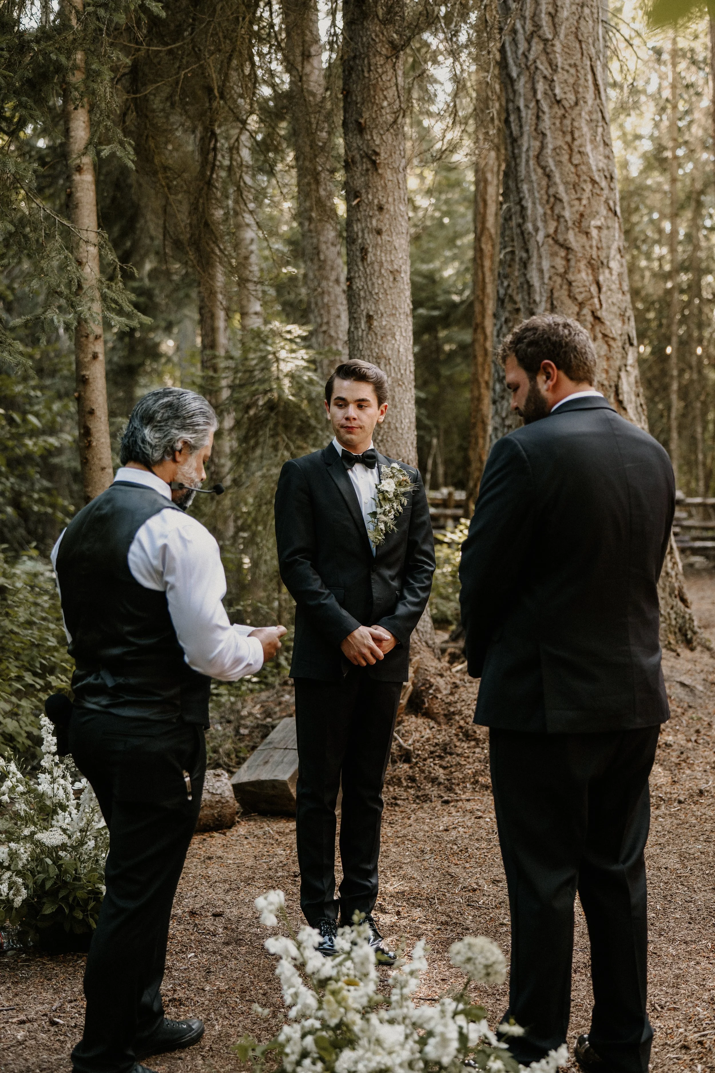 A wedding ceremony taking place outdoors in a forest with three men dressed in formal suits, two of them standing in front of a man with a pipe and a vest, amid trees and flowers.