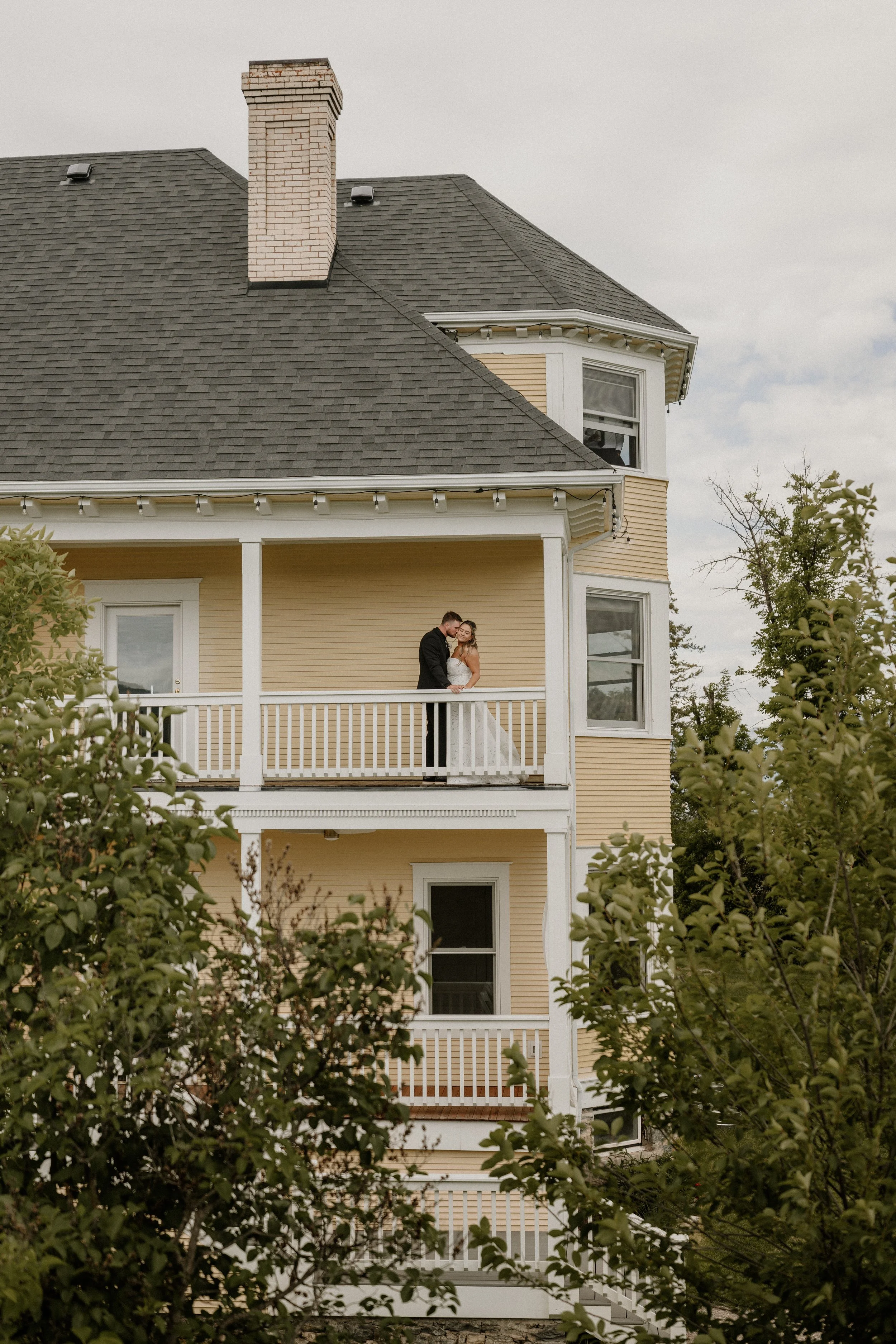 A yellow multi-story house with white trim and a porch is surrounded by green shrubs. A couple in wedding attire is standing on the porch, embracing.