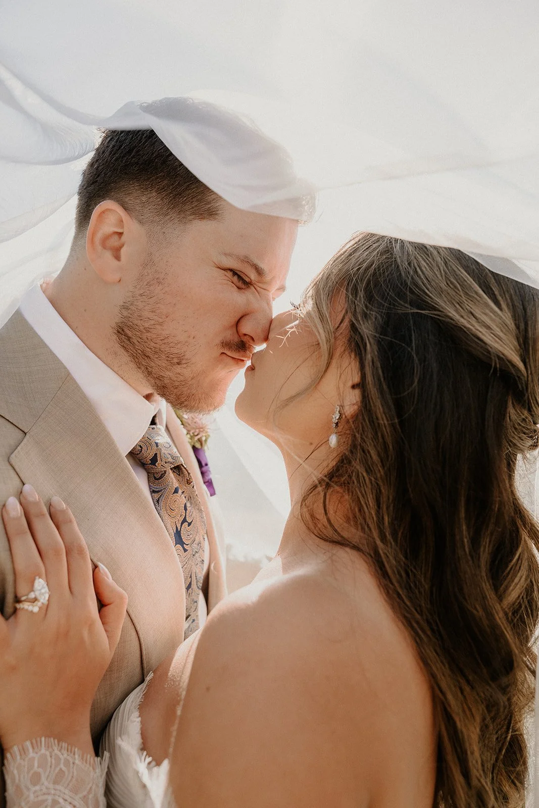 A close-up of a bride and groom about to kiss, under a veil, during their wedding. The groom is wearing a beige suit with a patterned tie, and the bride has long brown hair, earrings, and lace dress.