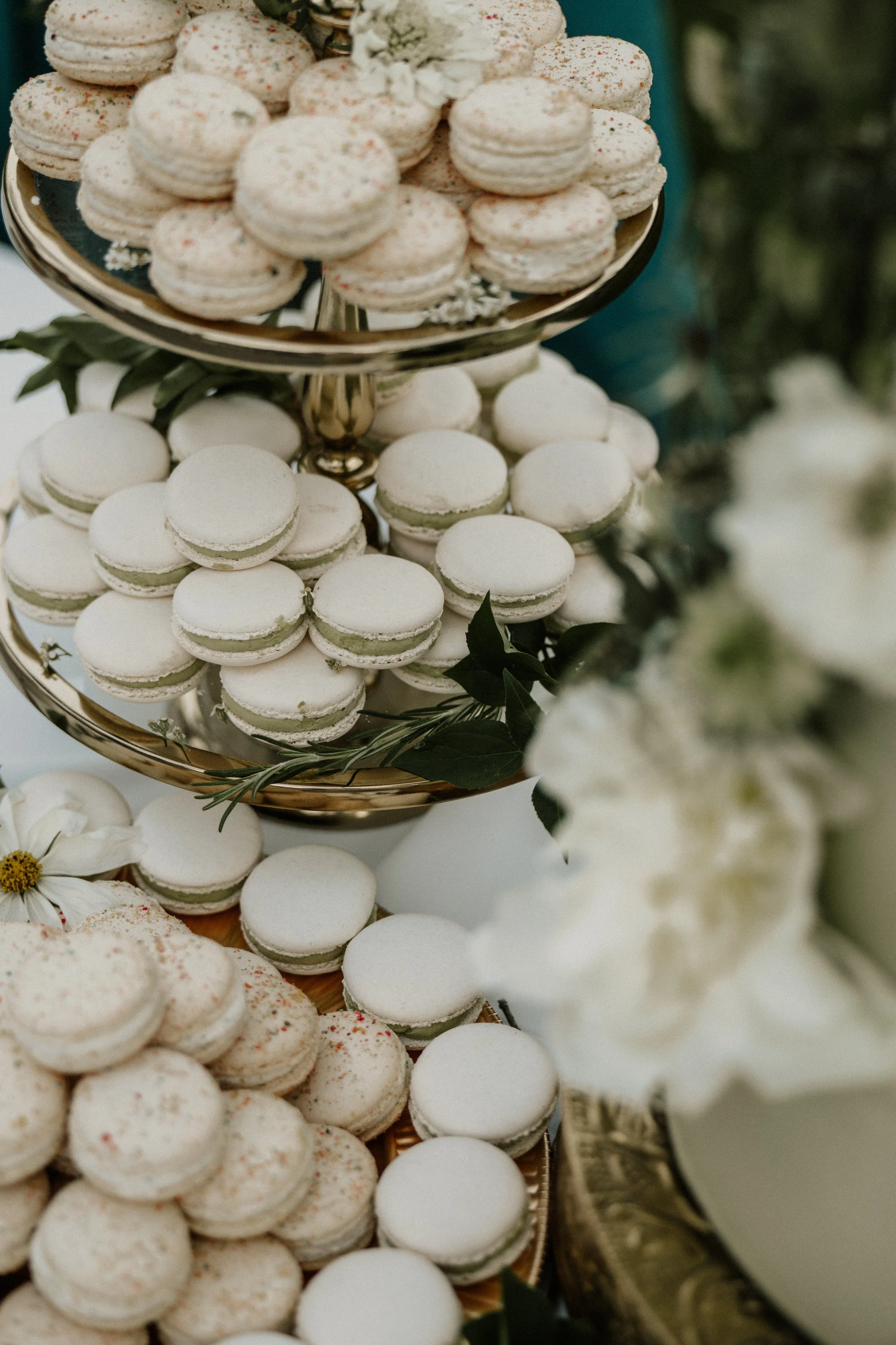 Close-up of a tiered tray with white macarons decorated with colorful sprinkles, garnished with greenery, and some on a decorative tray. Flowers are also visible in the scene.