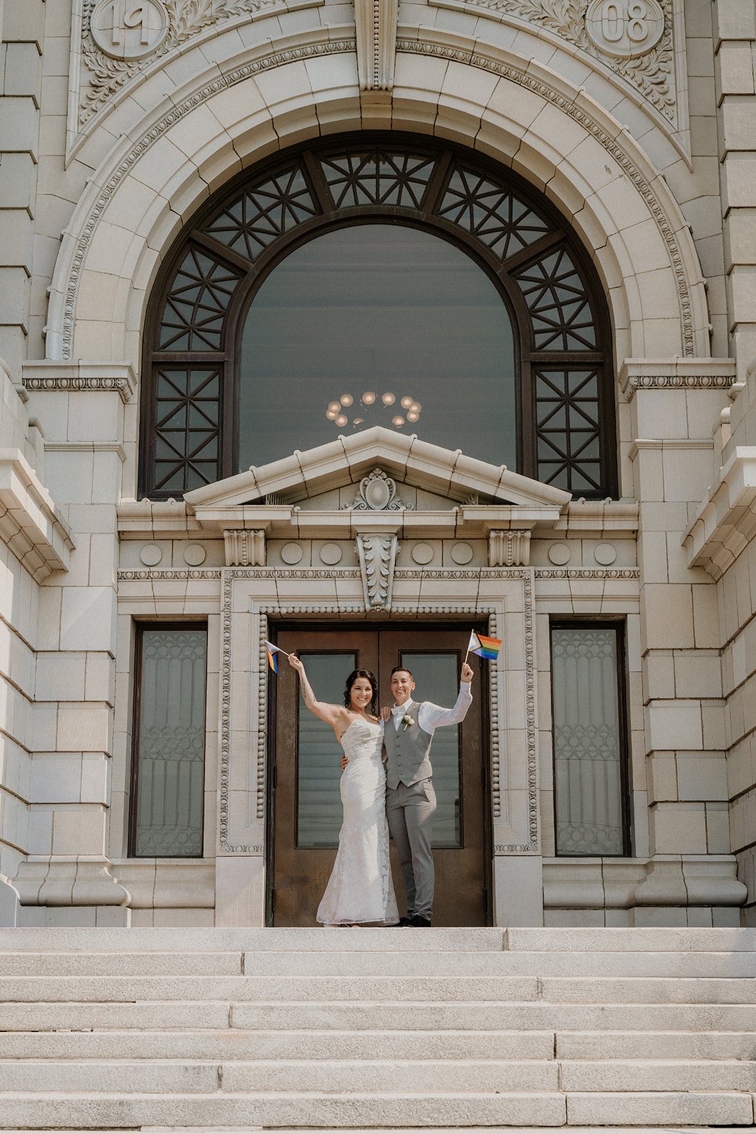 A newly married couple celebrating outside a historic building, holding rainbow pride flags, smiling, with stone steps and ornate architecture behind them.