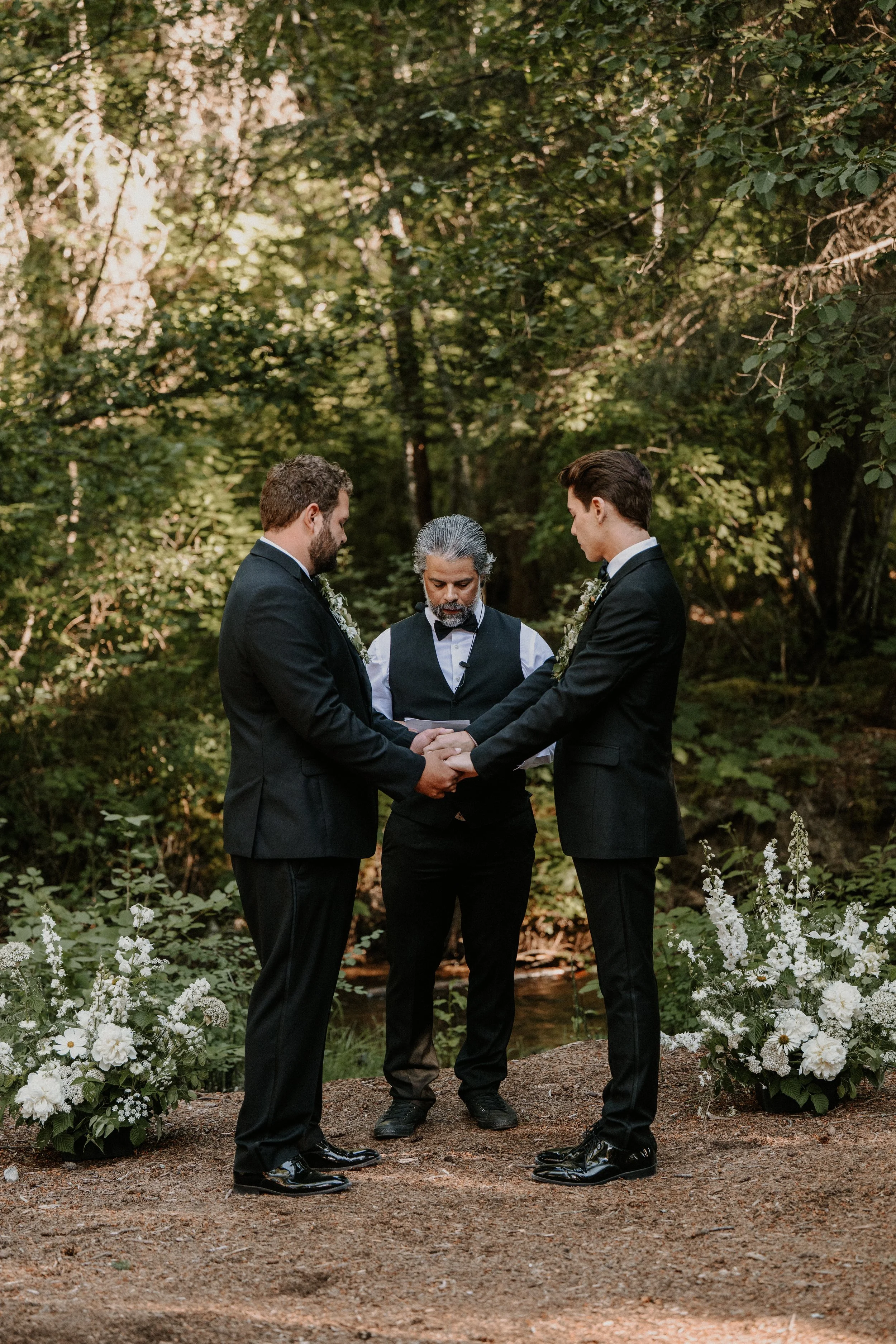 Two men in tuxedos holding hands during a wedding ceremony outdoors, with an officiant in a vest and bow tie, in a wooded area with floral arrangements.